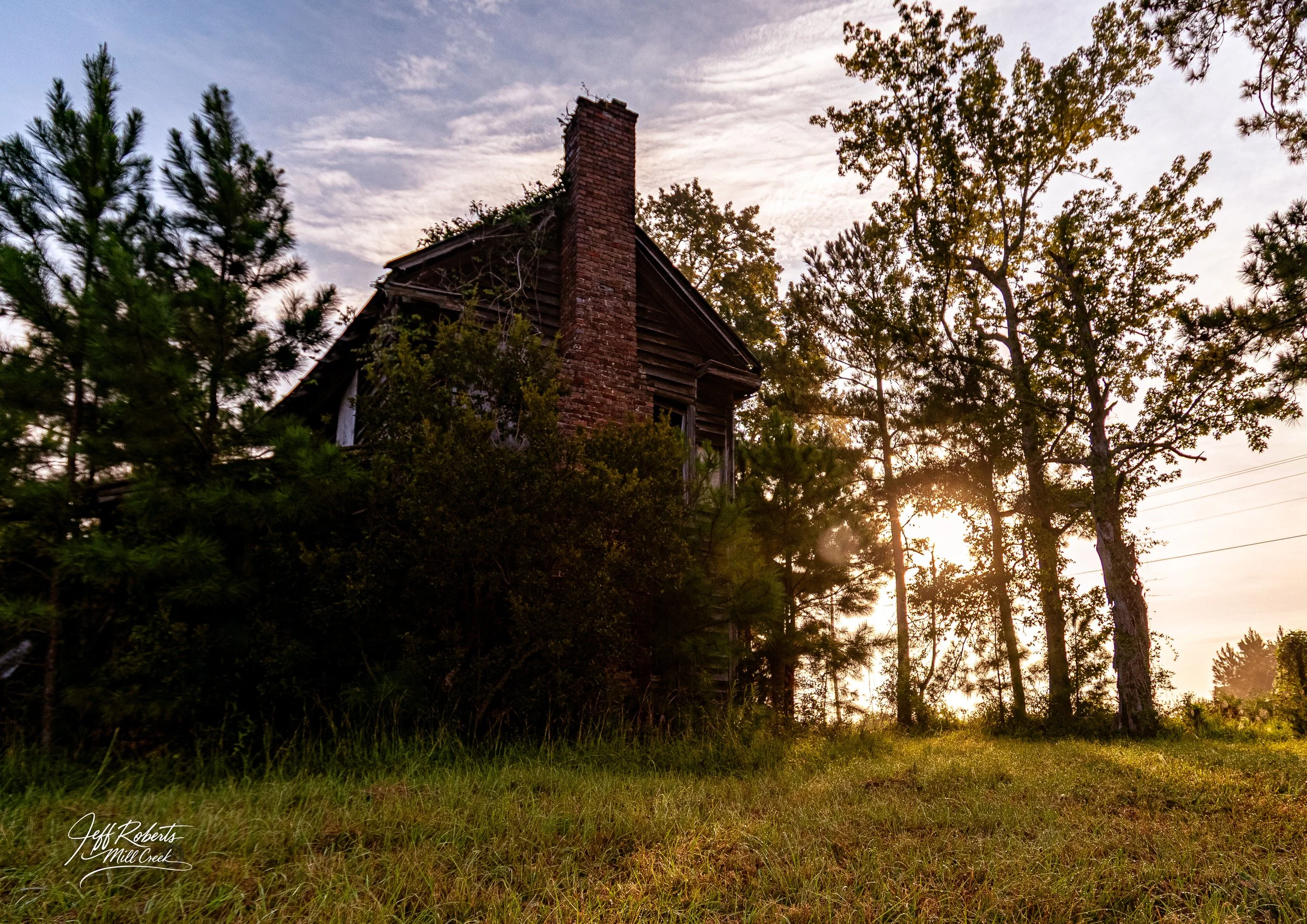 An abandoned house with a brick chimney, surrounded by trees and grass, at sunset with the sun low in the sky.