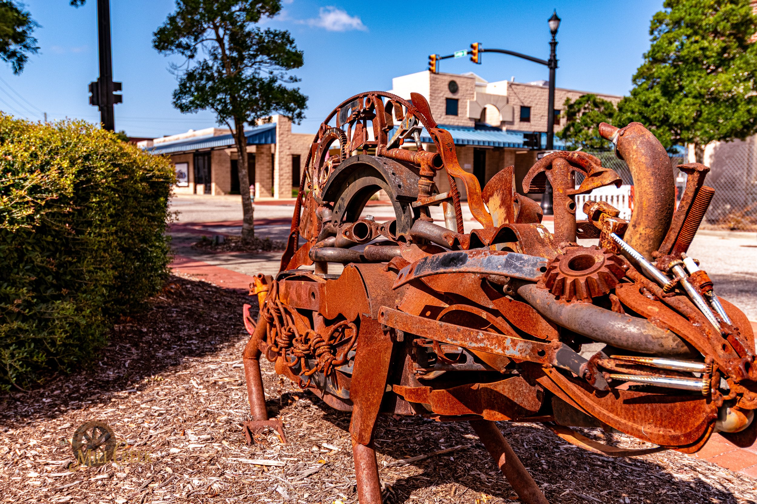 Sculpture of a horse made from rusted metal scrap in an outdoor park with trees and a building in the background.