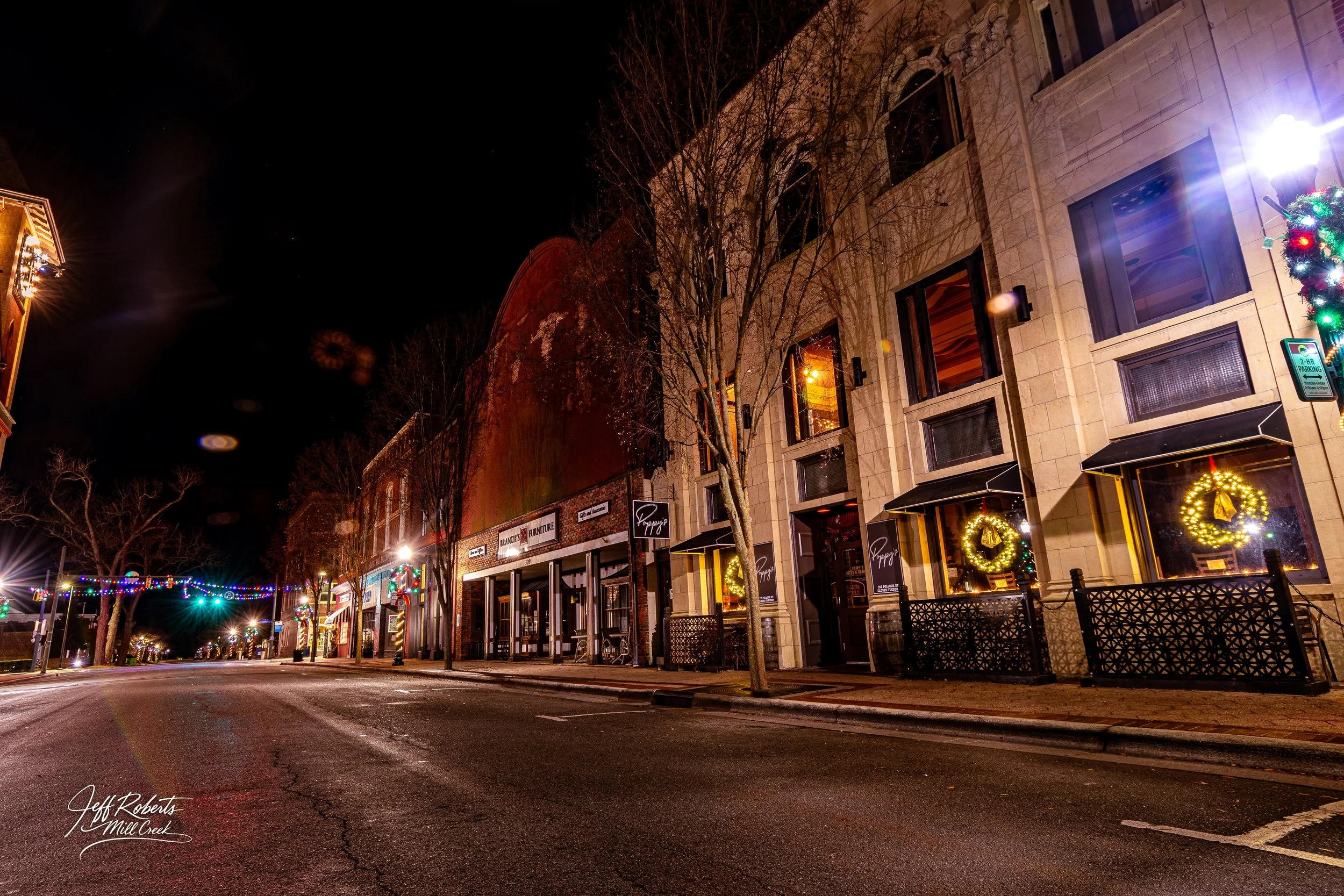 Empty downtown street decorated with Christmas wreaths and holiday lights at night, storefronts with awnings and warm yellow interior lights, leafless trees lining the sidewalk.
