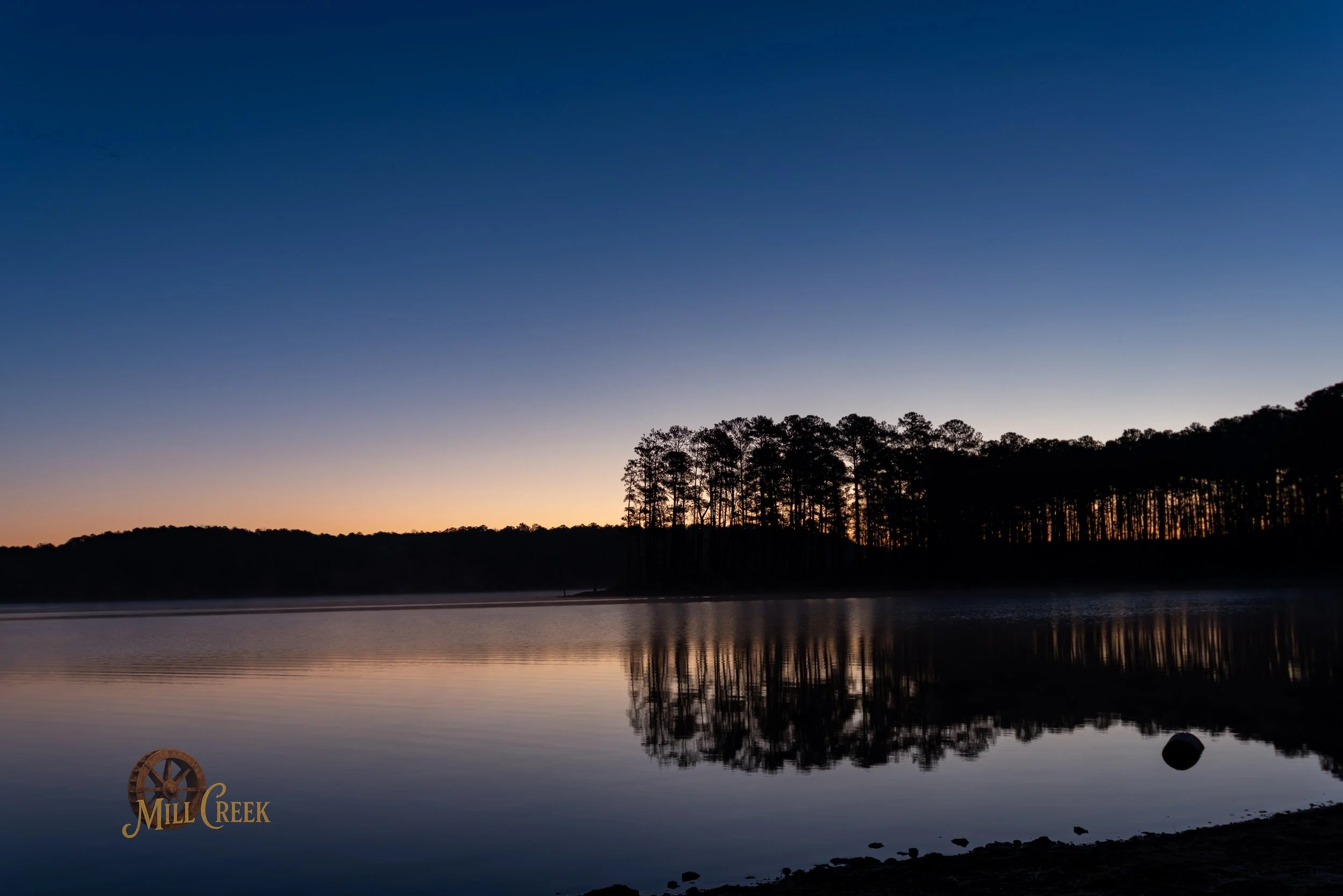 Sunset over a calm body of water with trees reflecting on the surface and the logo 'Mill Creek' at the bottom left corner.