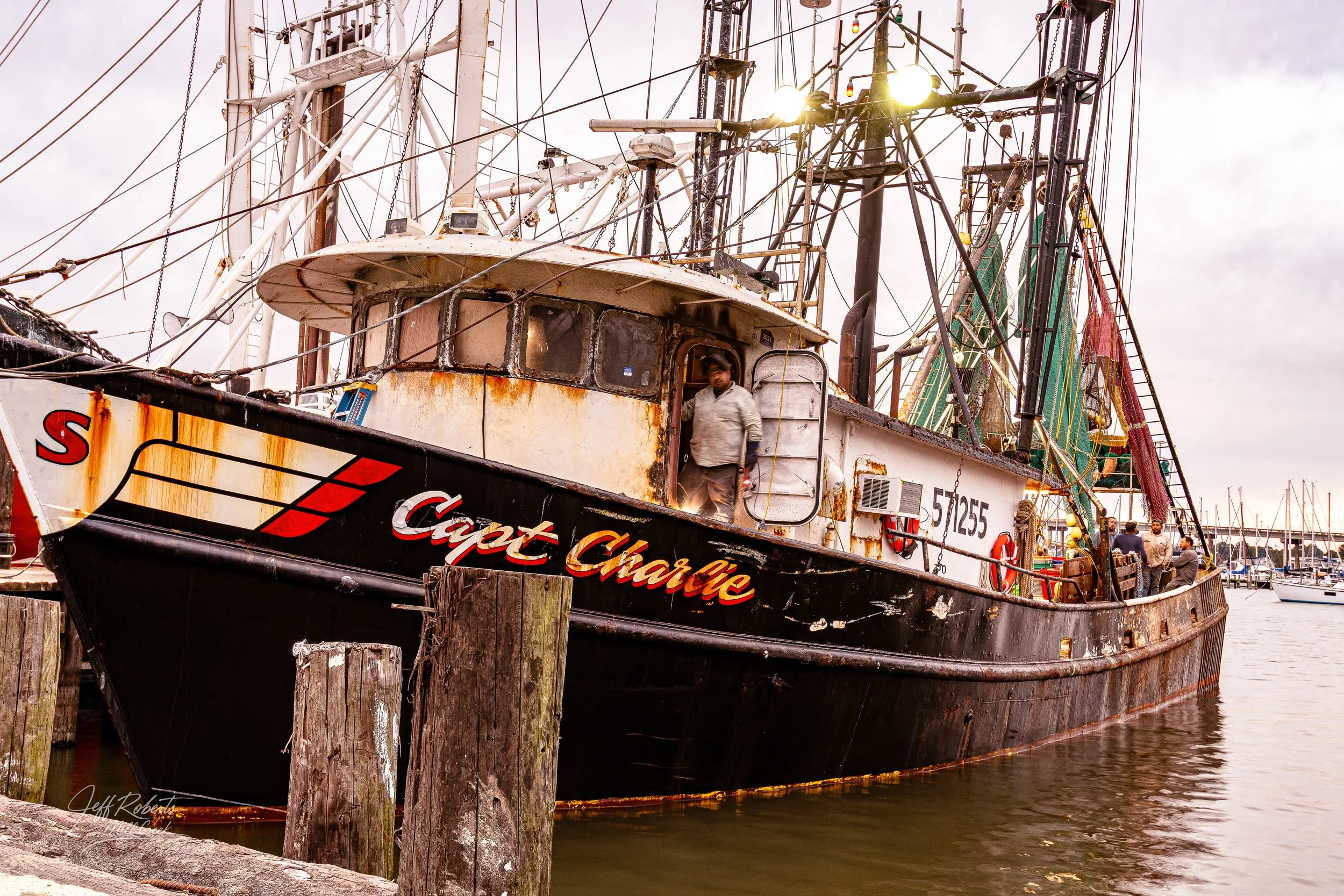 An old, rusty fishing boat named 'Captain Charlie' docked at a marina with several people on board. The boat has a weathered appearance with rust spots, a weathered superstructure, and various fishing equipment on deck. The marina has wooden pilings 