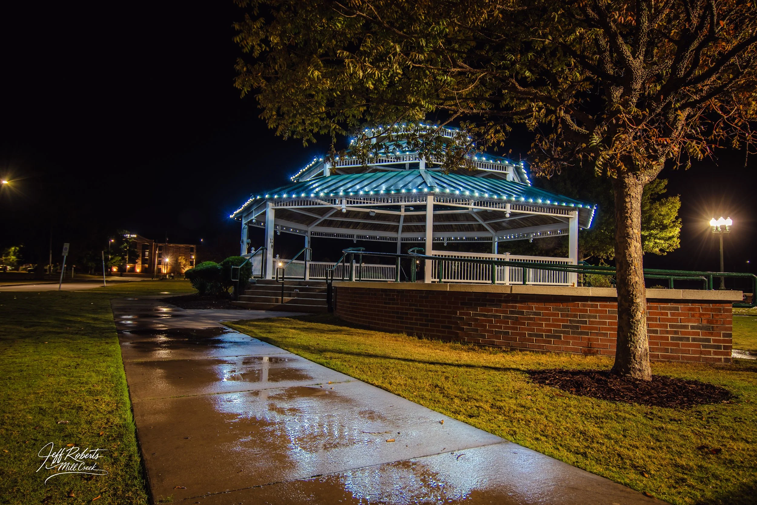 Nighttime view of a lit gazebo with a blue roof, surrounded by trees, with wet pavement reflecting the lights and an illuminated streetlamp in the background.