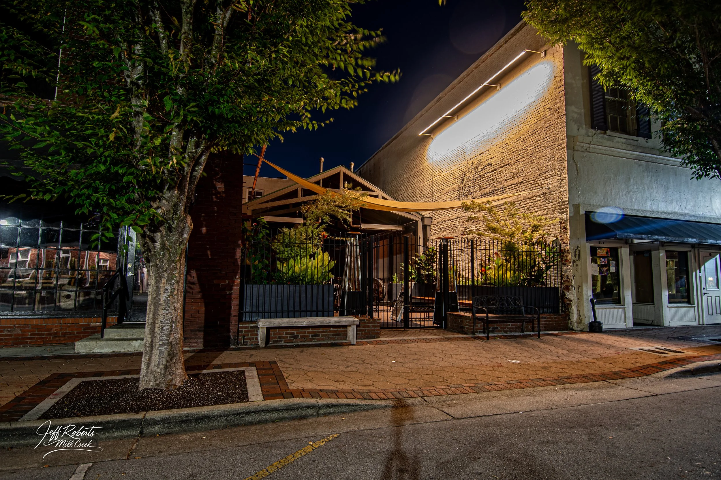Nighttime street view showing a building with a brick and concrete facade, outdoor seating, greenery, and a large illuminated sign on the wall.
