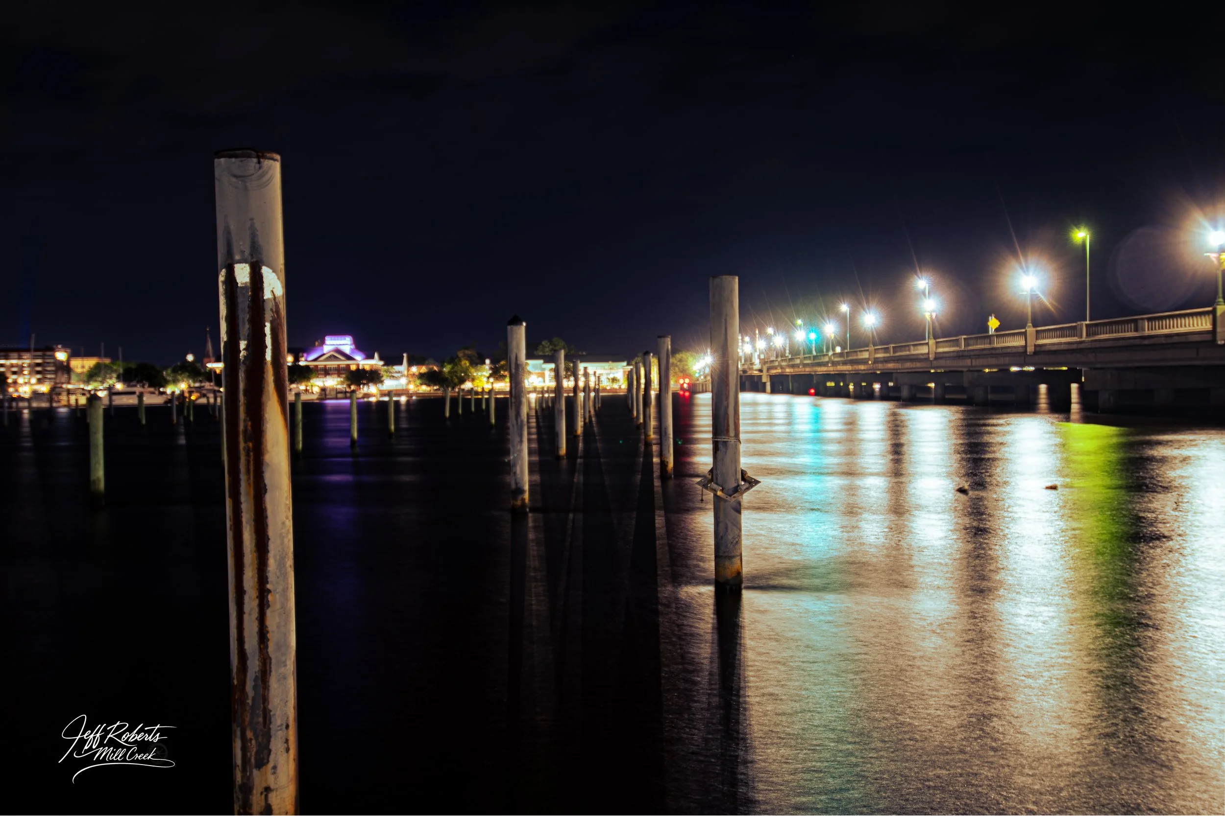 Nighttime view of a river with old rusty posts sticking out of the water, illuminated city lights in the background, and a bridge with bright streetlights reflected on the water.