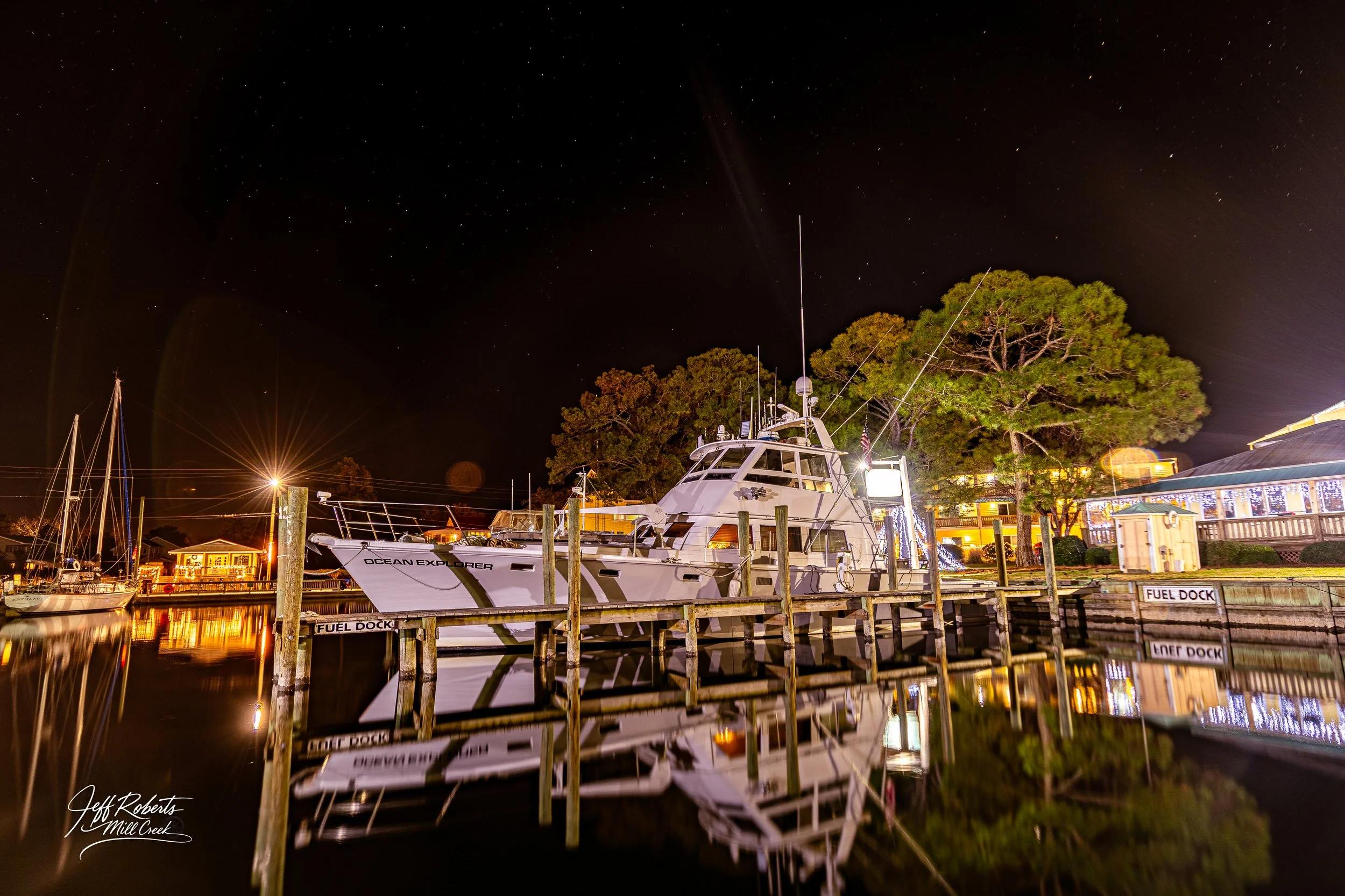 Nighttime scene of a white yacht named 'Ocean Explorer' docked at a pier with calm water reflecting the boat and surrounding lights. There are trees and buildings with holiday lights in the background, and the sky is filled with stars.