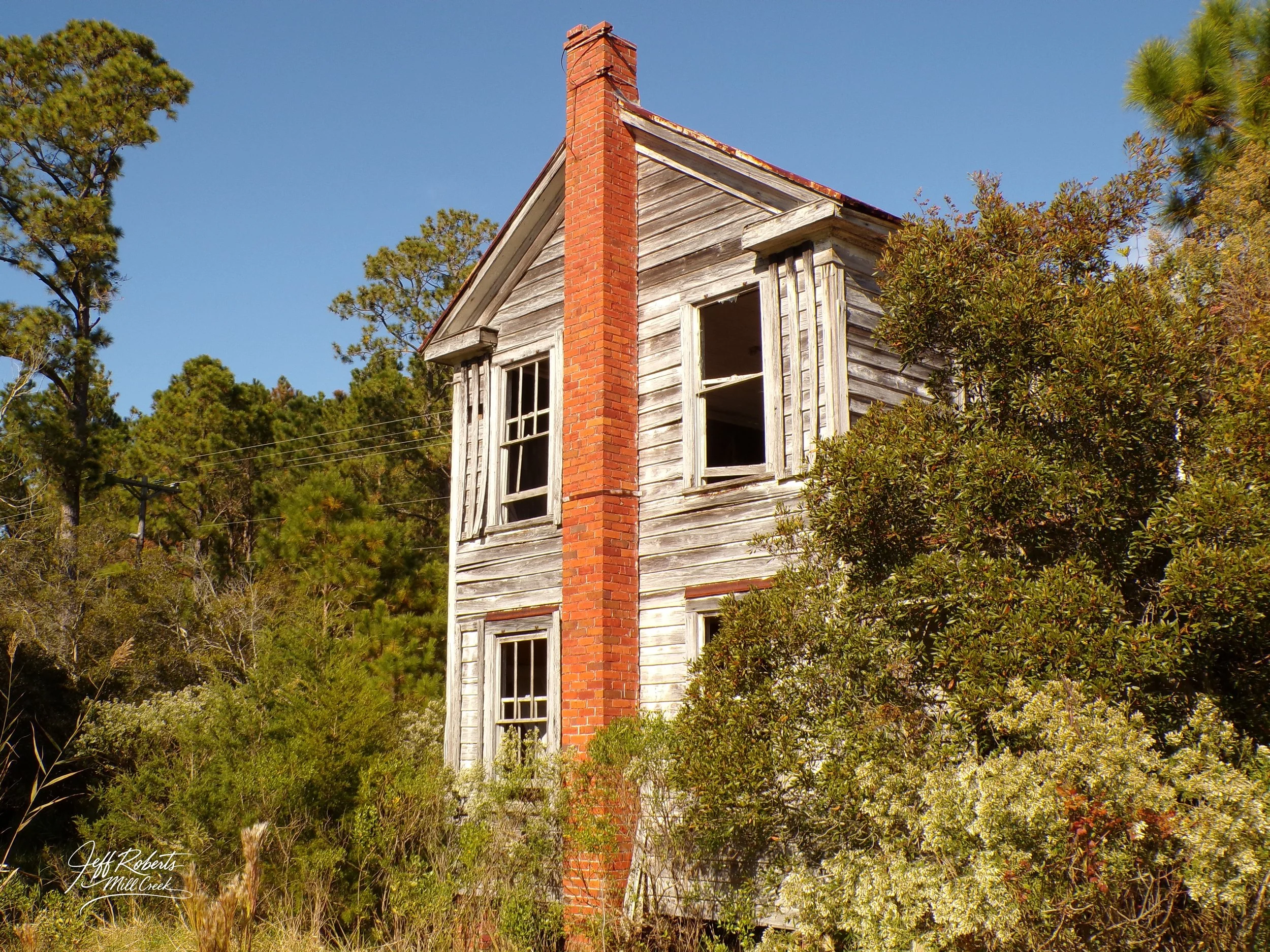 An old, abandoned two-story wooden house with peeling paint, broken windows, and a brick chimney, surrounded by overgrown bushes and trees under a clear blue sky.