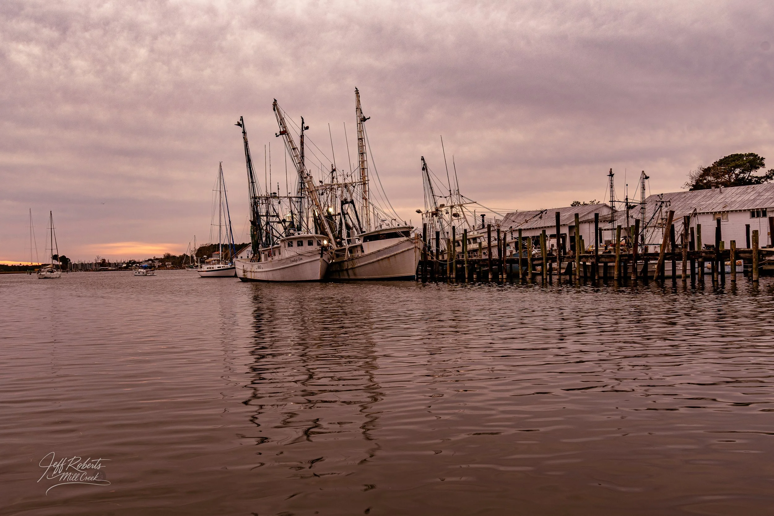 Boats docked at a pier along a calm river, with a cloudy sky overhead, during sunset.
