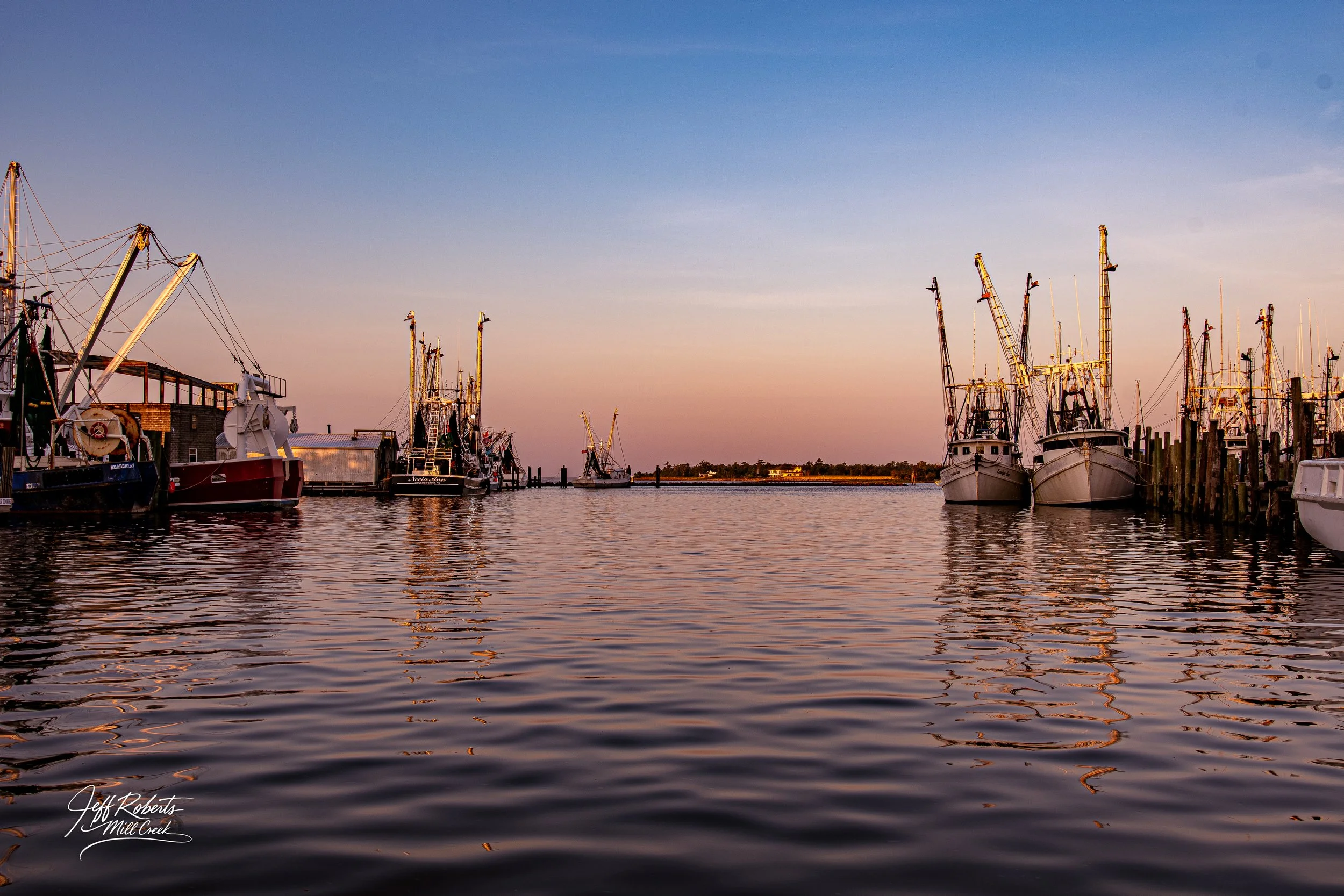 Fishing boats docked at a marina during sunset with calm water reflecting the boats and sky.