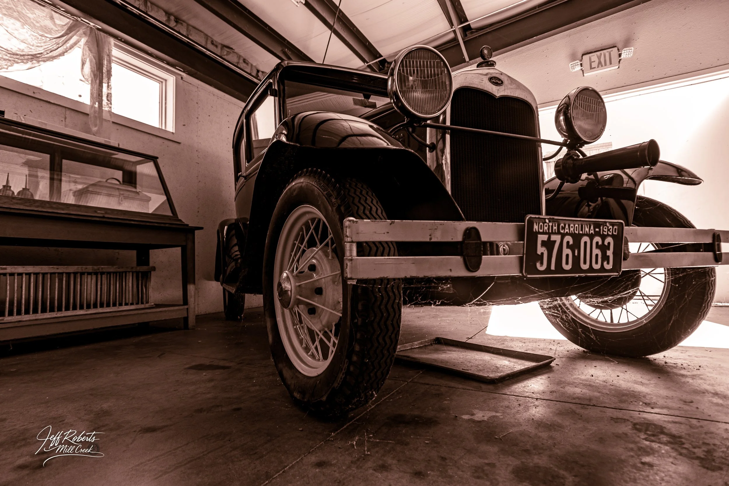 A vintage black and white car with a North Carolina license plate from 1930 is parked indoors, with cobwebs on the front bumper and wheels. The car has large round headlights, a prominent grille, and a spare tire mounted on the side. Nearby, there is