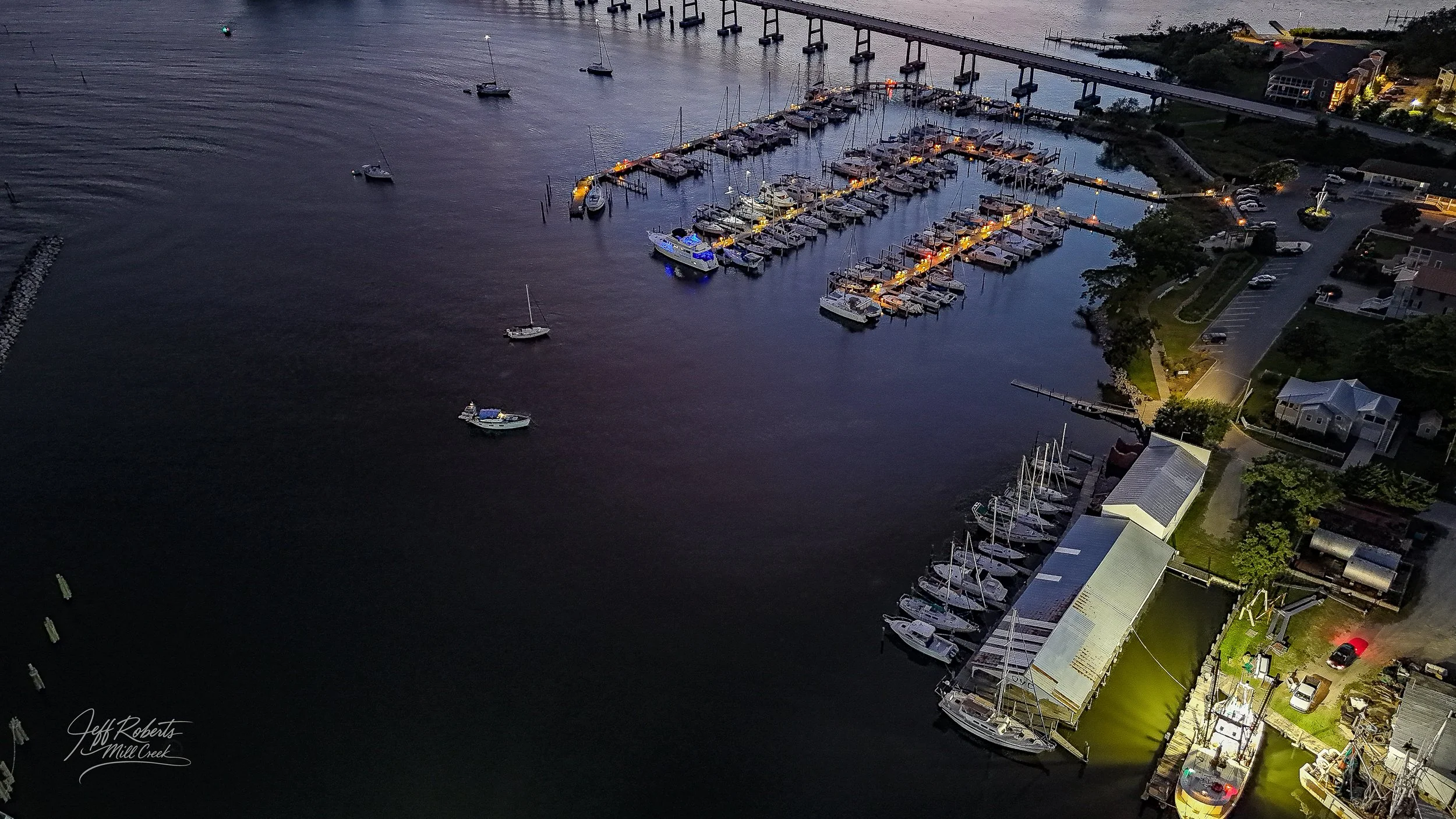 An aerial view of a marina at dusk, with several boats docked and some sailing in the water. The marina is illuminated with lights, and nearby residential buildings and parking lot are visible.