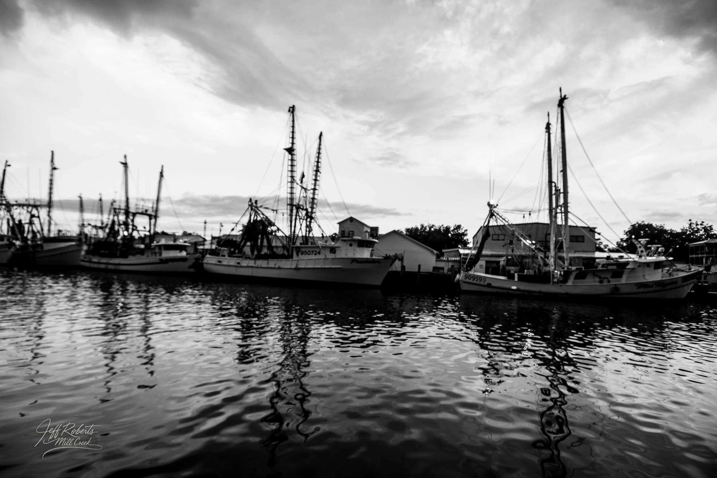 Black and white photo of several boats docked at a pier with buildings and trees in the background under a cloudy sky.