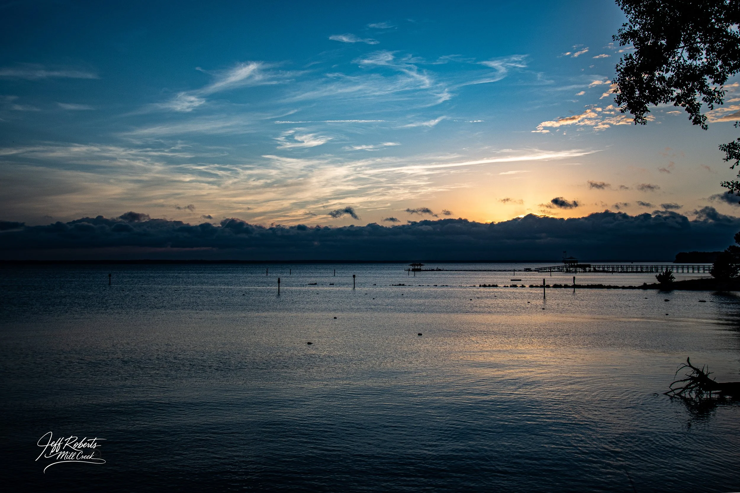Sunset over a calm body of water with a pier extending into the distance and trees on the right side.