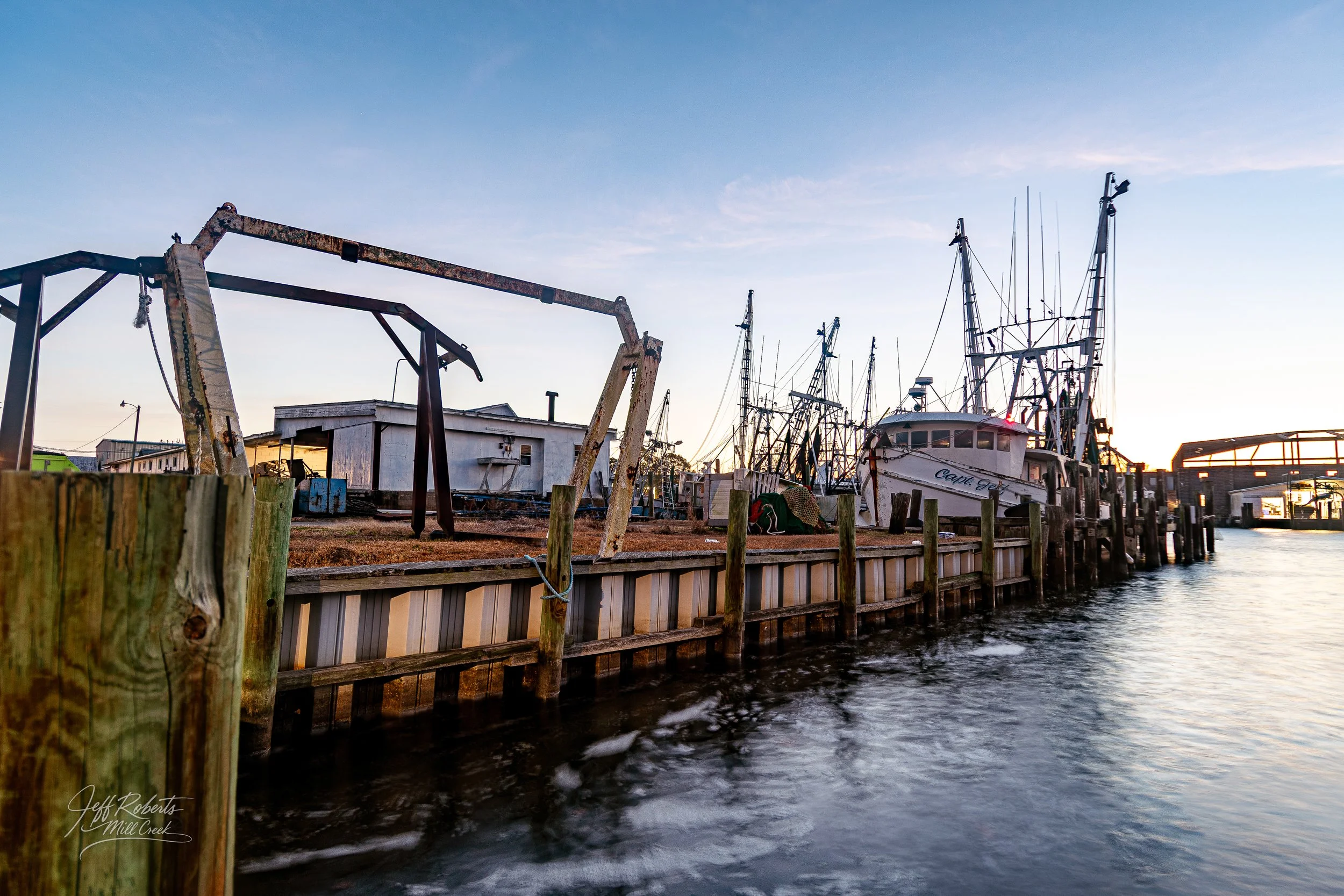 View of a marina at sunset with boats docked, wooden posts, and a waterway in the foreground.