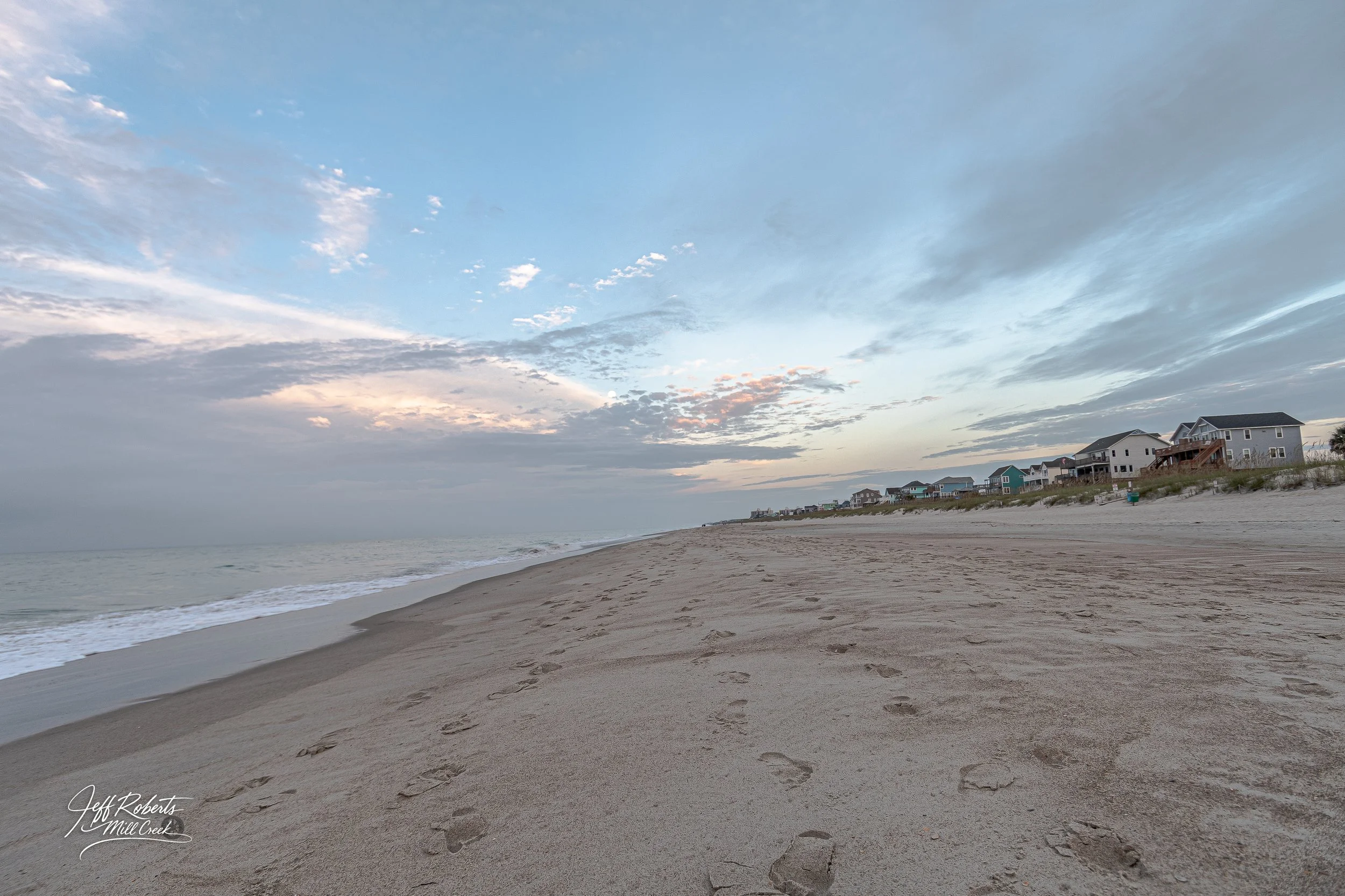 Empty beach with footprints in the sand, houses in the distance, and a cloudy sky during sunset.