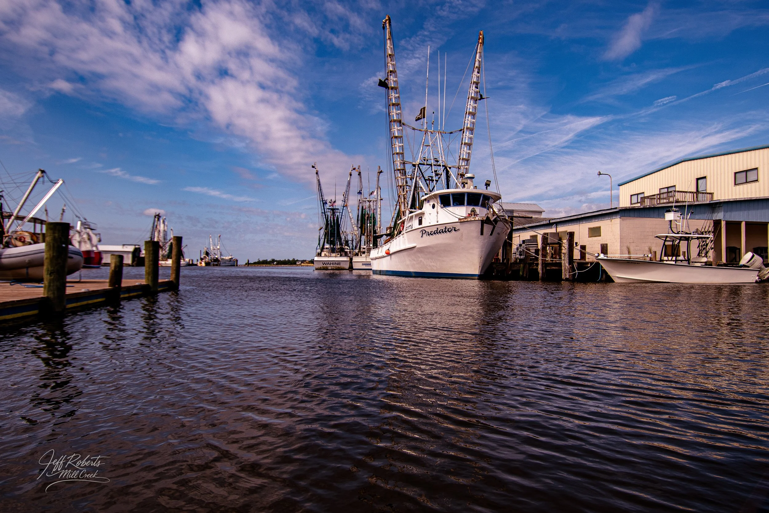 Marina with boats docked at the pier, including a large boat named Predator, under a blue sky with scattered clouds.
