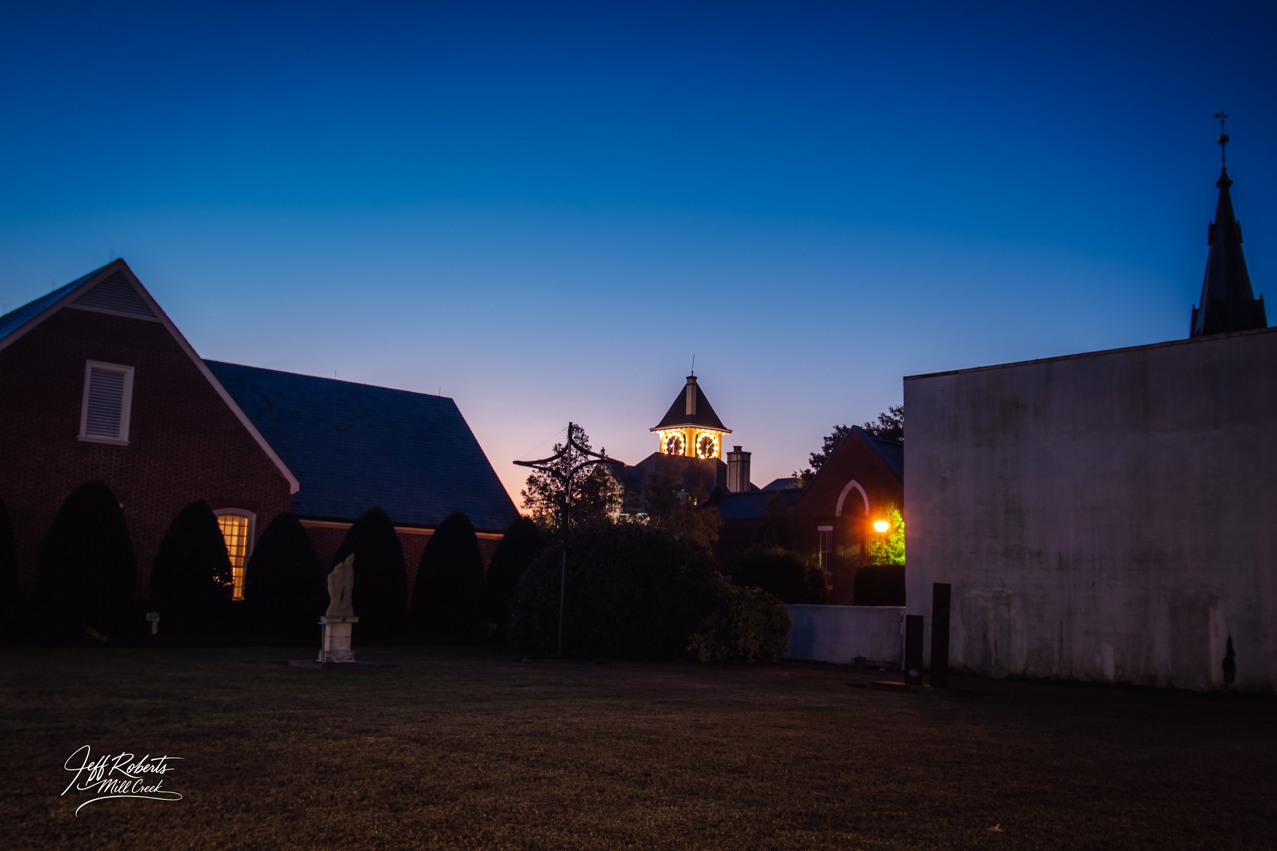Evening view of a church with a lit clock tower, houses, and a statue, under a clear blue sky, in a small town.