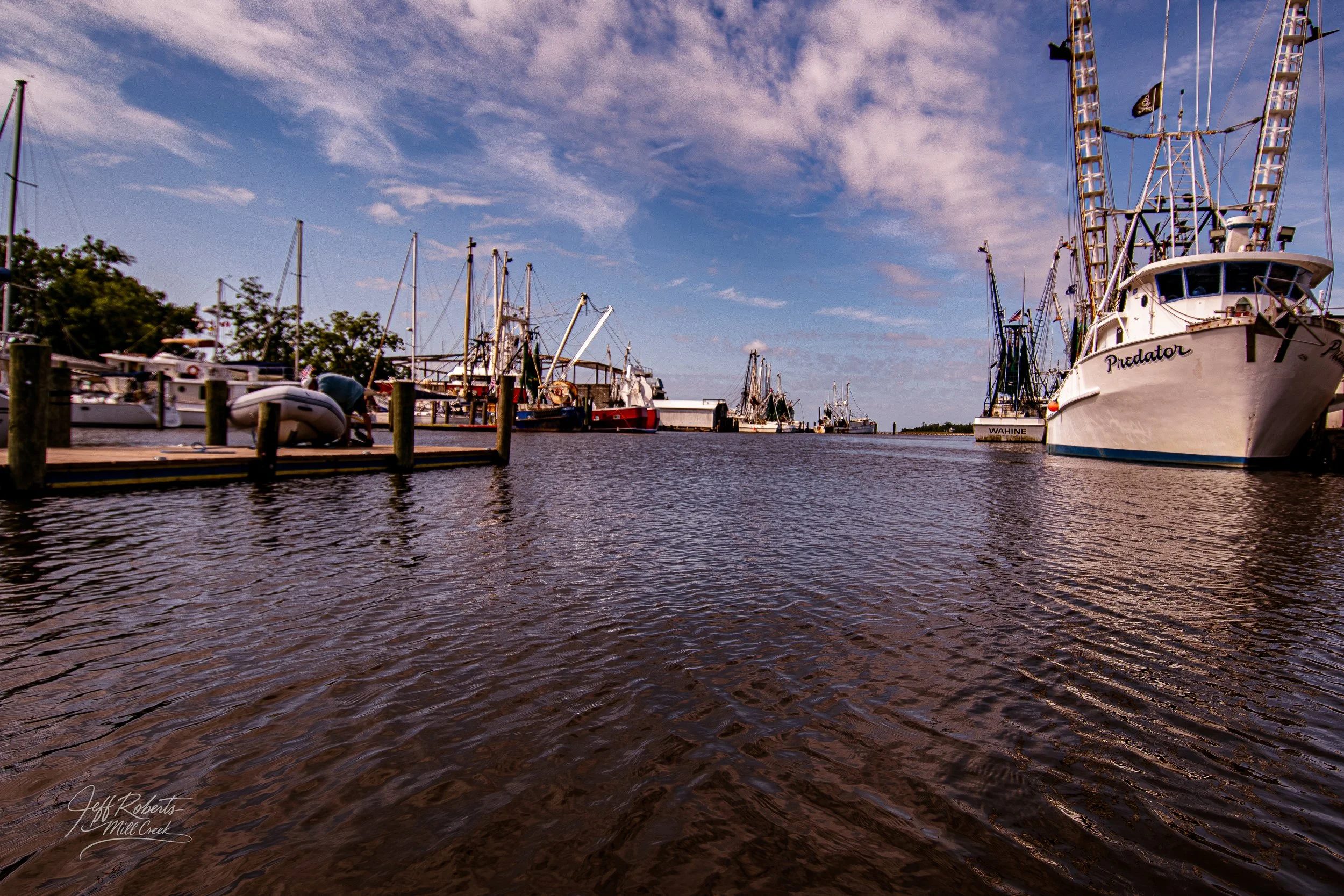 Marina with boats docked along the pier, including various sailboats and fishing boats, under a partly cloudy sky.