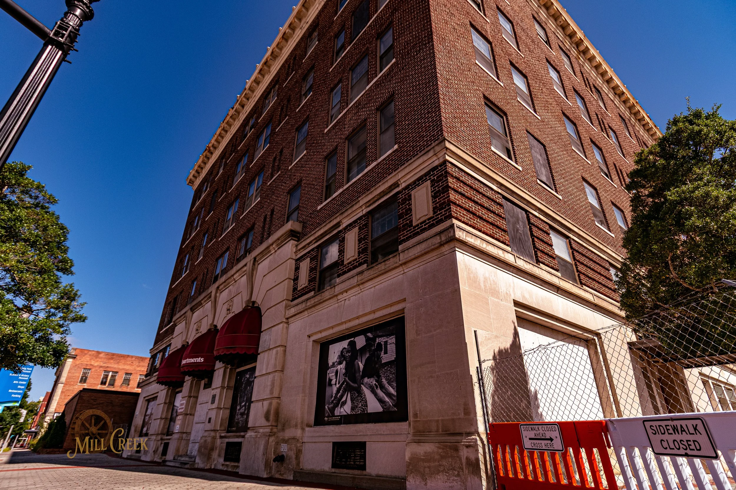 A tall brick building with multiple windows and a stone base, with street signs and a construction barrier in the foreground.