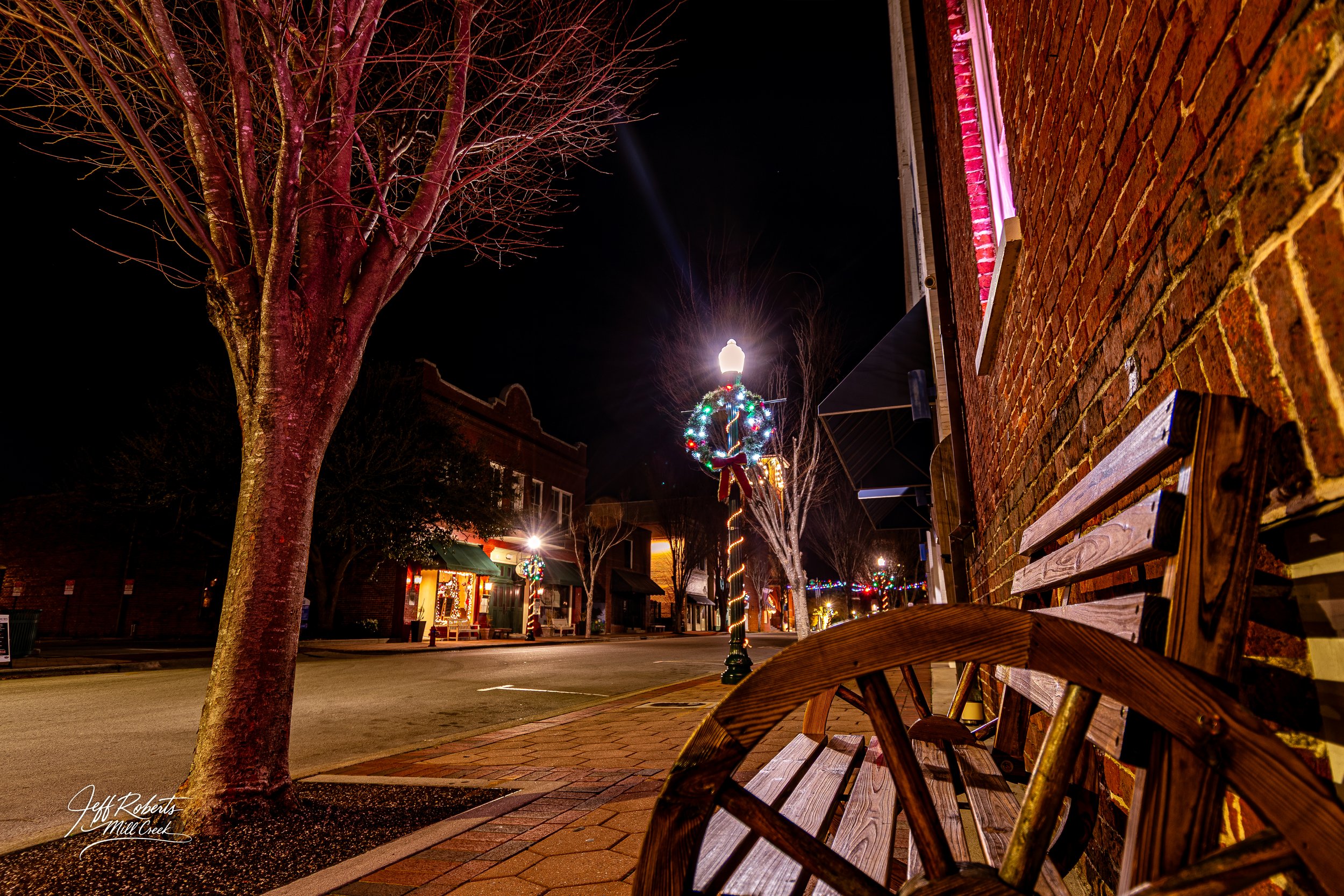 Empty wooden bench on brick sidewalk, decorated street lights, and lit storefronts on a quiet street at night with Christmas decorations.