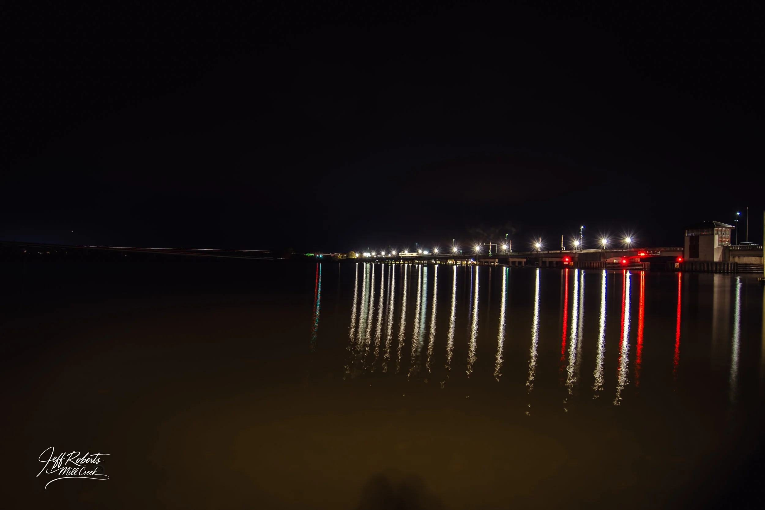 Nighttime view of a pier with bright lights reflected on calm water, and a building at the far right.