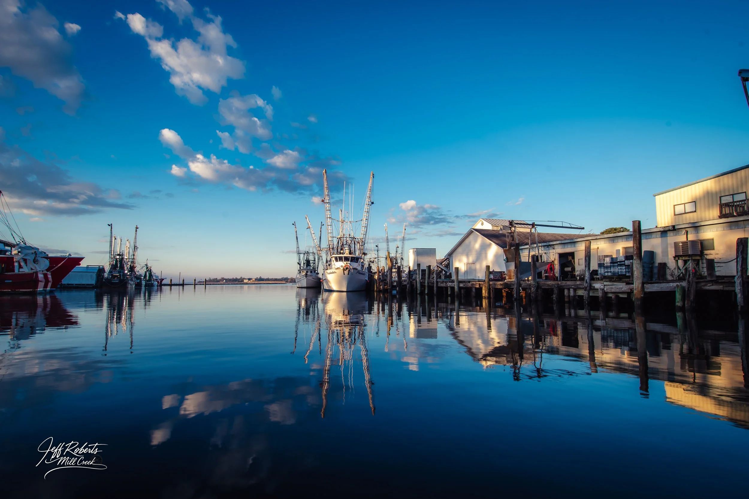 Boats docked along a wooden pier on a calm body of water under a partly cloudy sky during sunset.