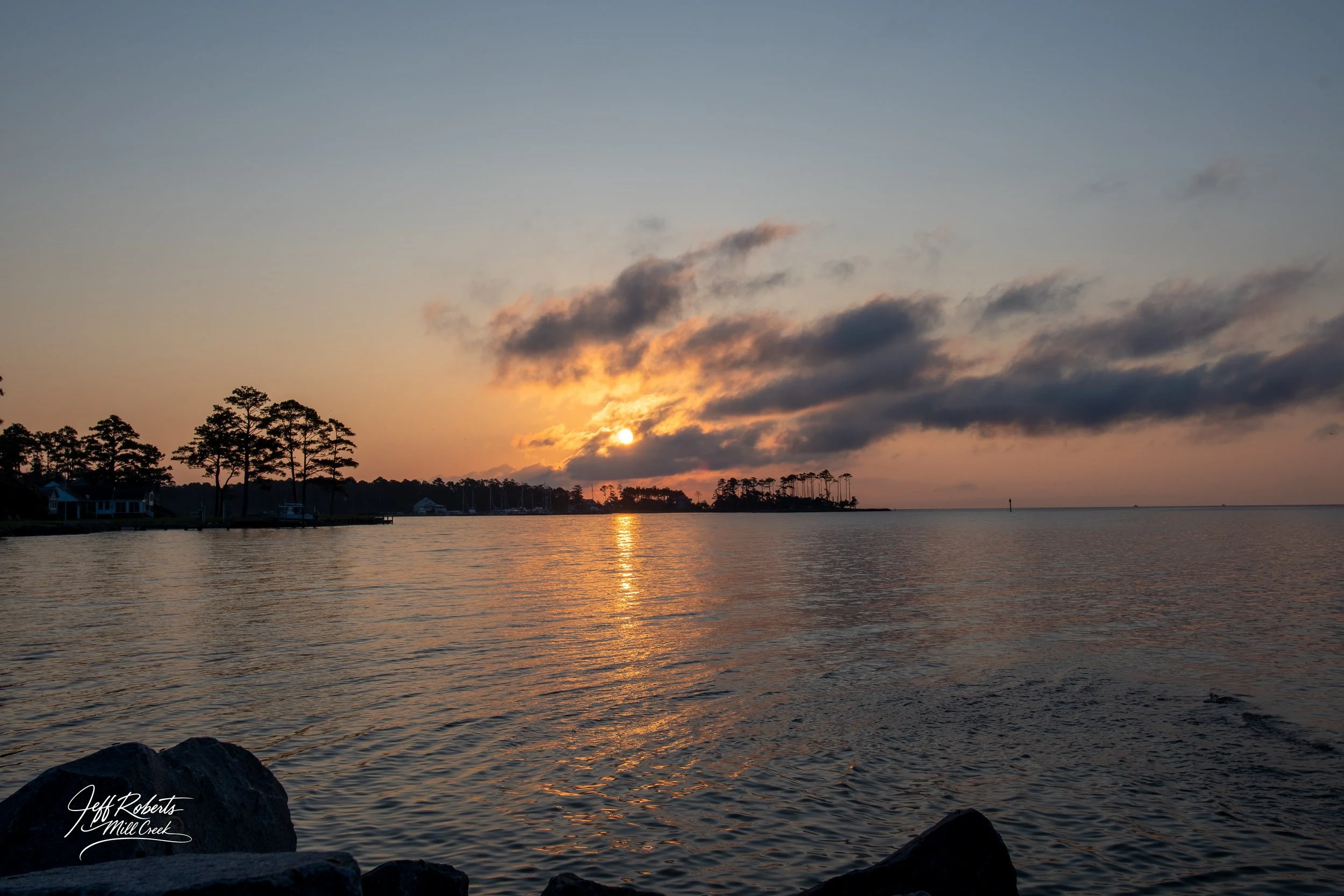 Sunset over the water with silhouettes of trees and houses along the shoreline, reflecting light on the surface of the water.