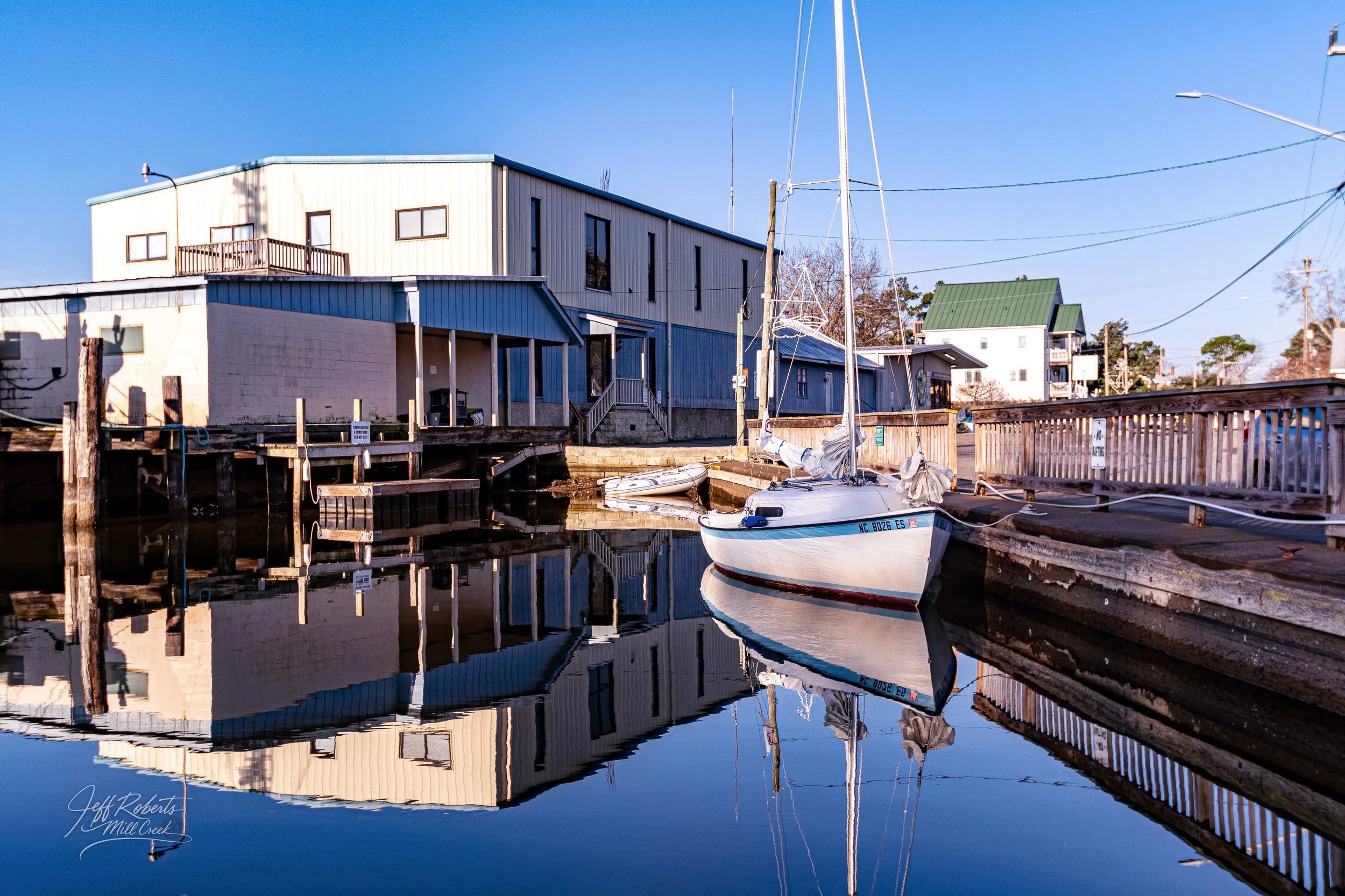 A small sailboat docked in a calm waterway with buildings, a wooden dock, and power lines in the background, reflecting on the water in clear weather.