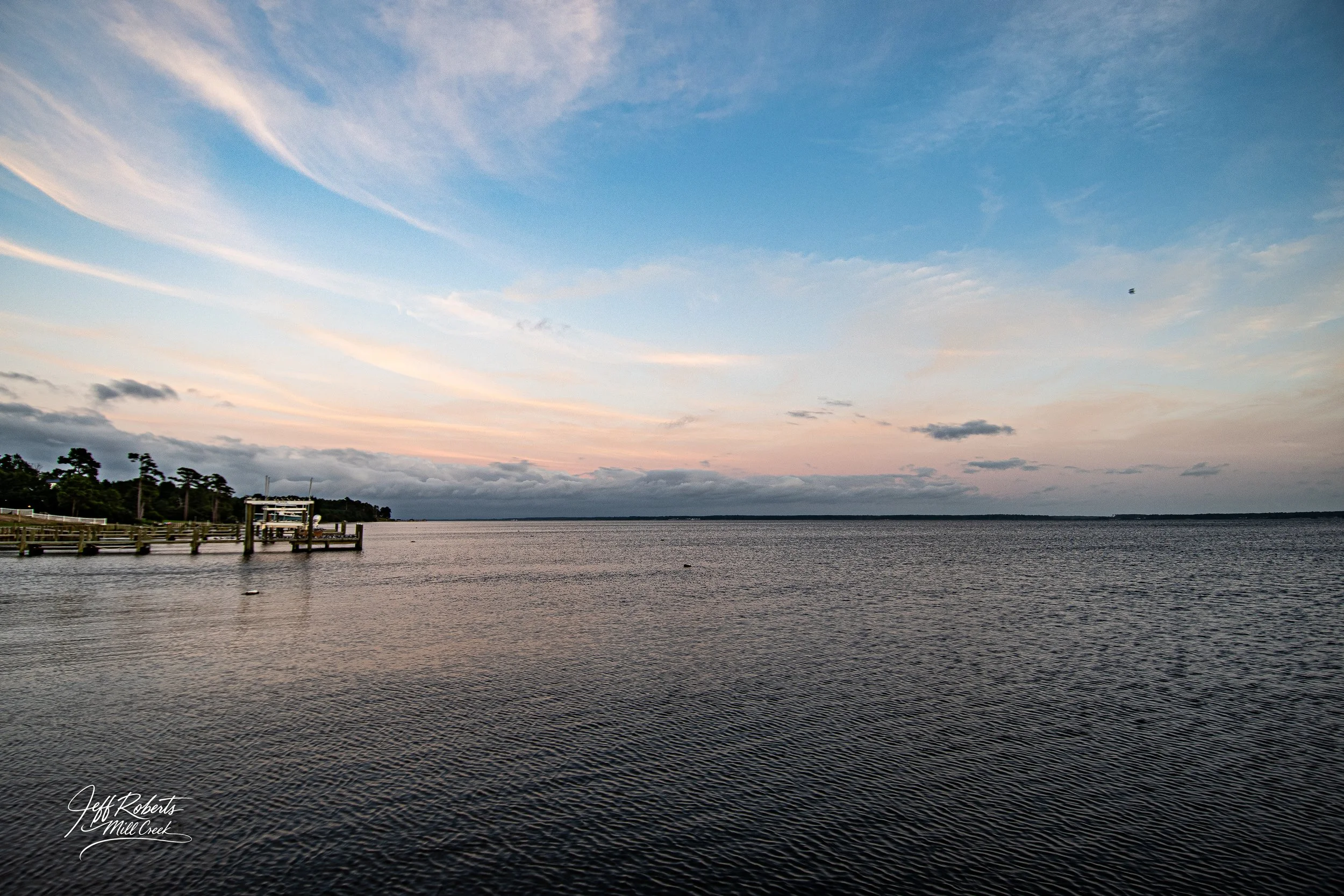A peaceful body of water with a dock on the left side, under a partly cloudy sky during sunset or sunrise.