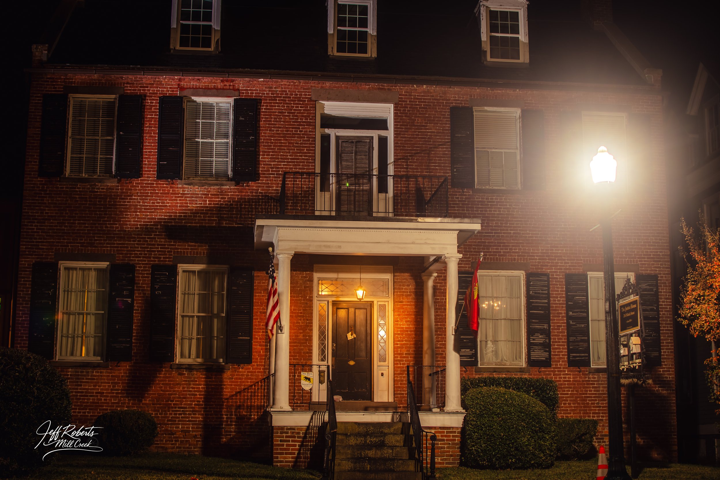 A brick building at night with illuminated windows, a front door with a small porch, two flags near the entrance, bushes on either side, a street lamp shining from the right side, and a sign on the right side of the building.