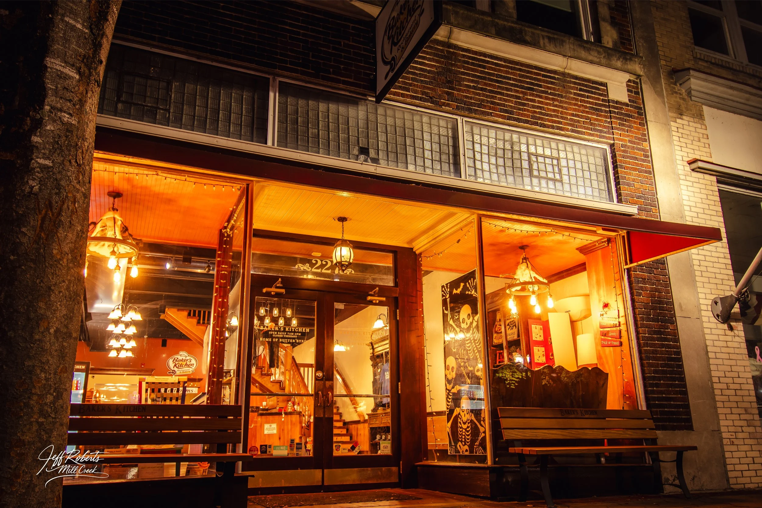 Night view of a warmly lit restaurant with glass doors, wooden benches outside, Halloween decorations including skeletons and pumpkins, and a staircase visible inside.