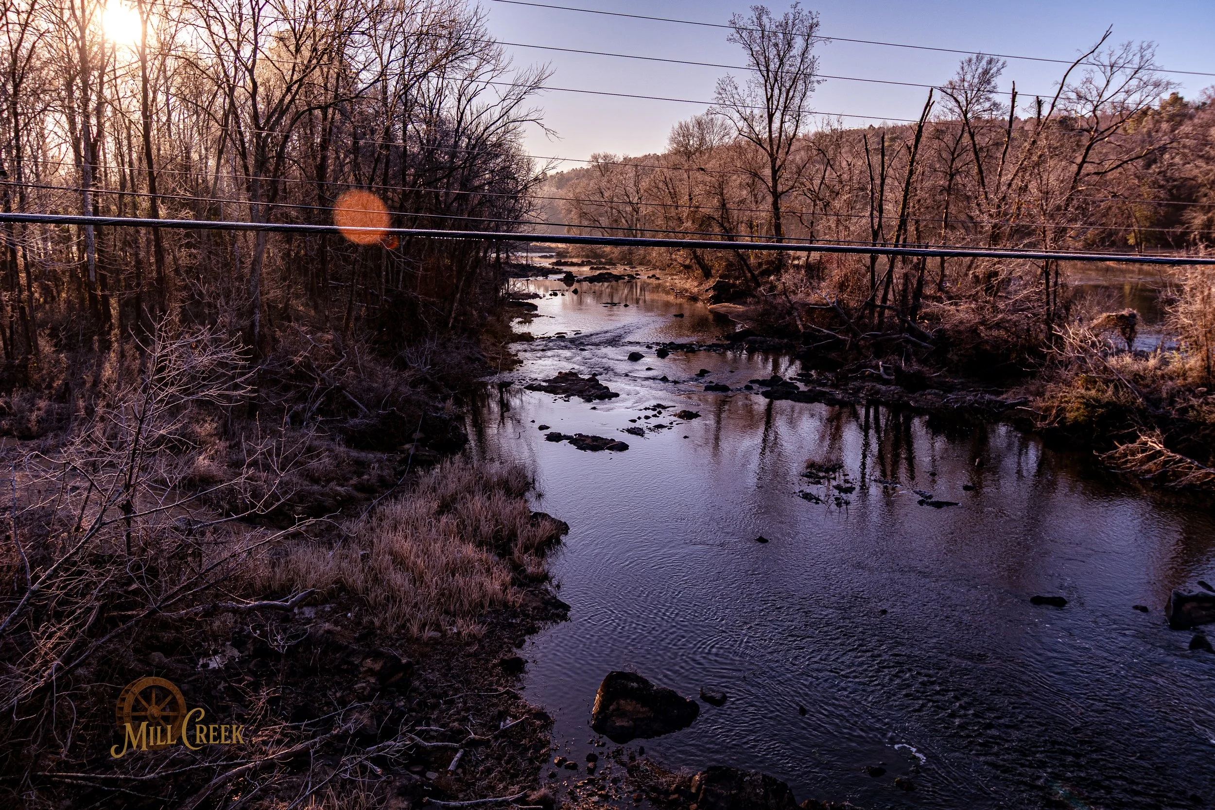 A river flowing through a wooded landscape during sunset, with leafless trees on both sides and power lines crossing above.