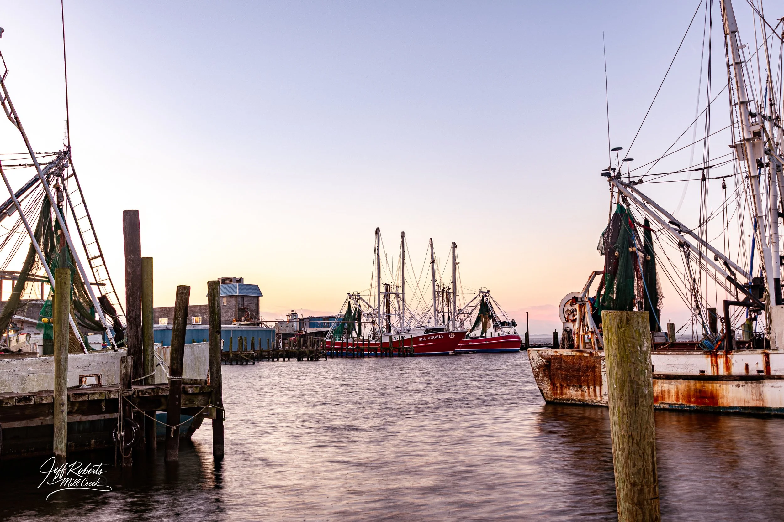 View of a harbor at sunset with sailboats docked at the pier, some boats with rusted hulls, and several wooden posts in the water.