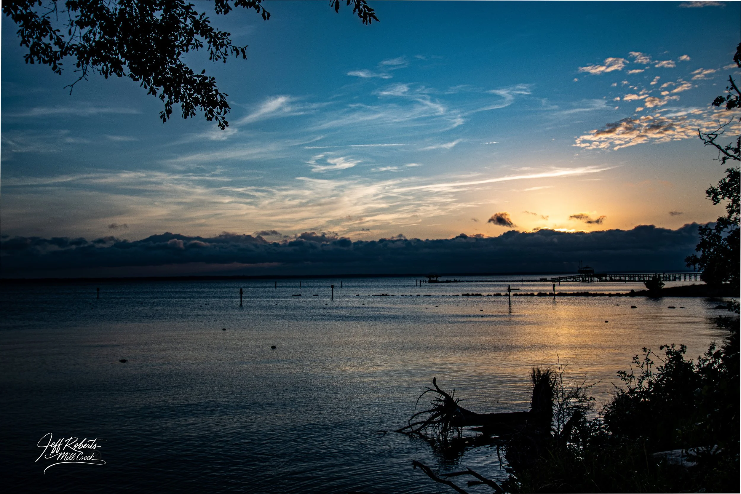 Sunset over a calm body of water, with a pier extending into the distance, dark clouds on the horizon, and branches framing the scene at the top and bottom.