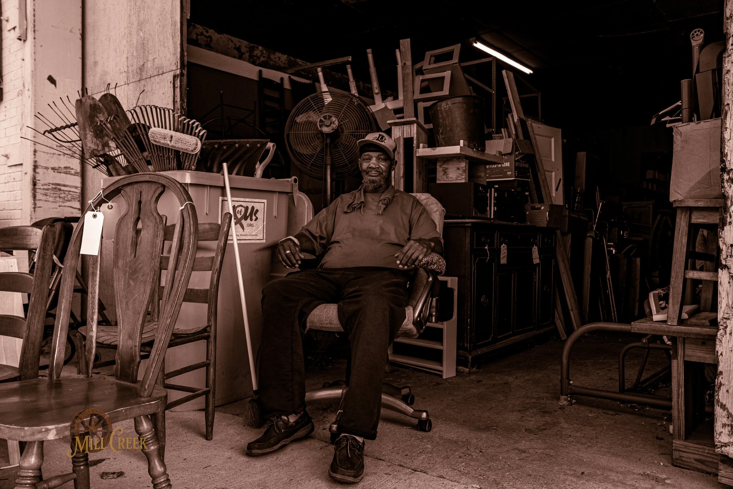 A man sitting in a cluttered room, surrounded by furniture and various household items.