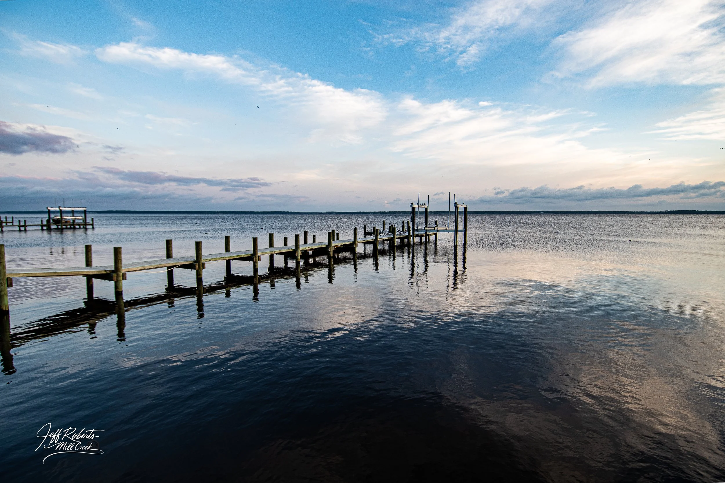 A wooden pier extending into a calm body of water under a partly cloudy sky during dusk or dawn.