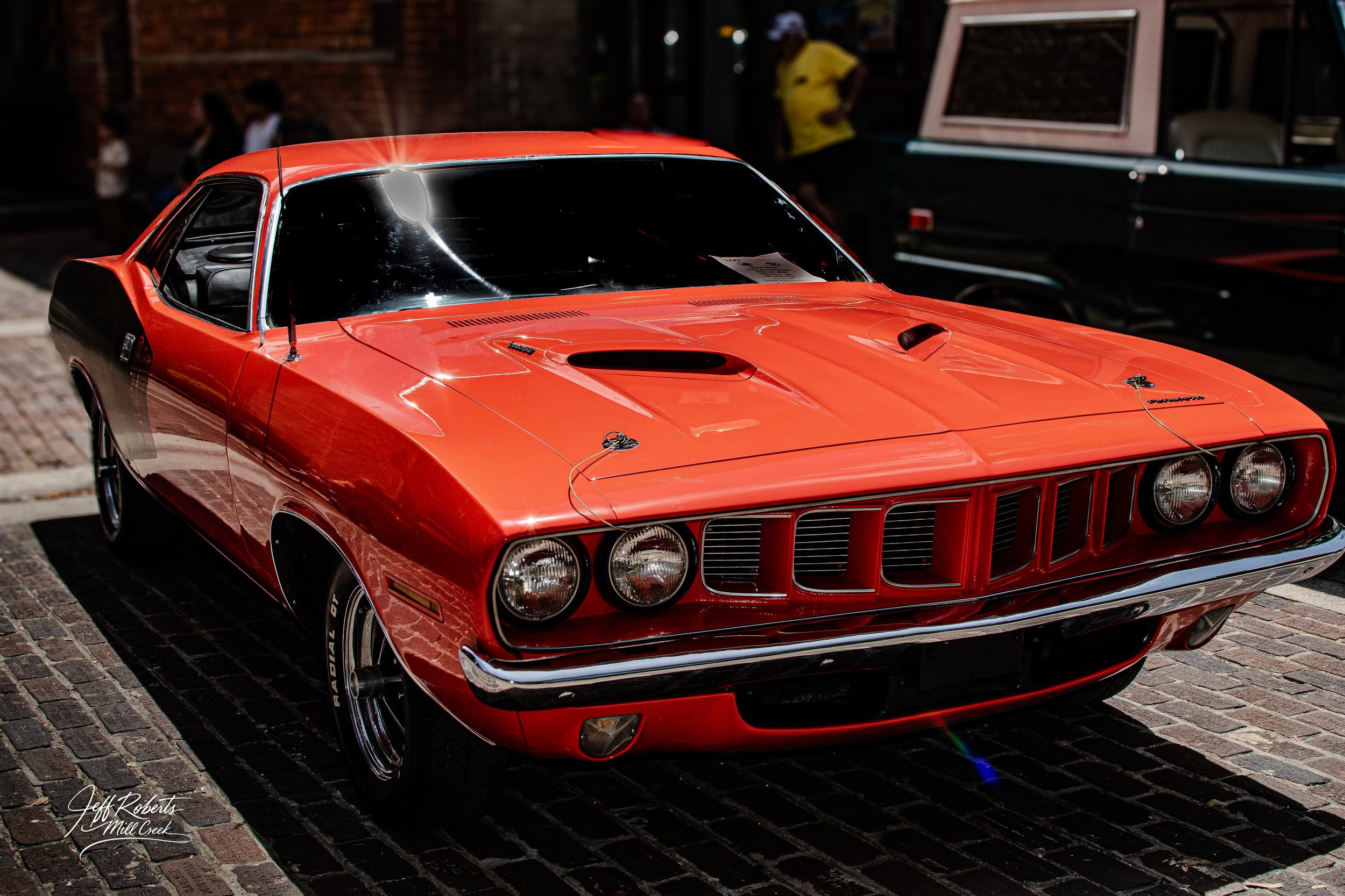A vintage orange muscle car with a black hood and black side accents parked on a brick street at night, reflecting lights on its shiny surface.