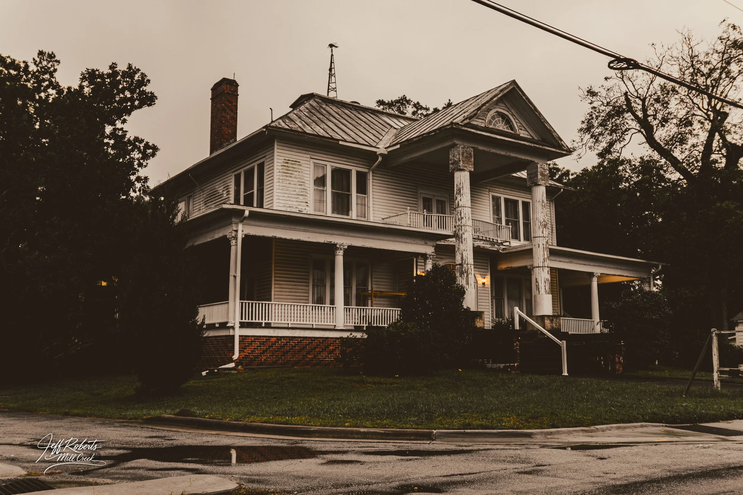An old two-story house with peeling white paint, large columns, a brick chimney, and a wraparound porch, surrounded by trees and grass, under an overcast sky.