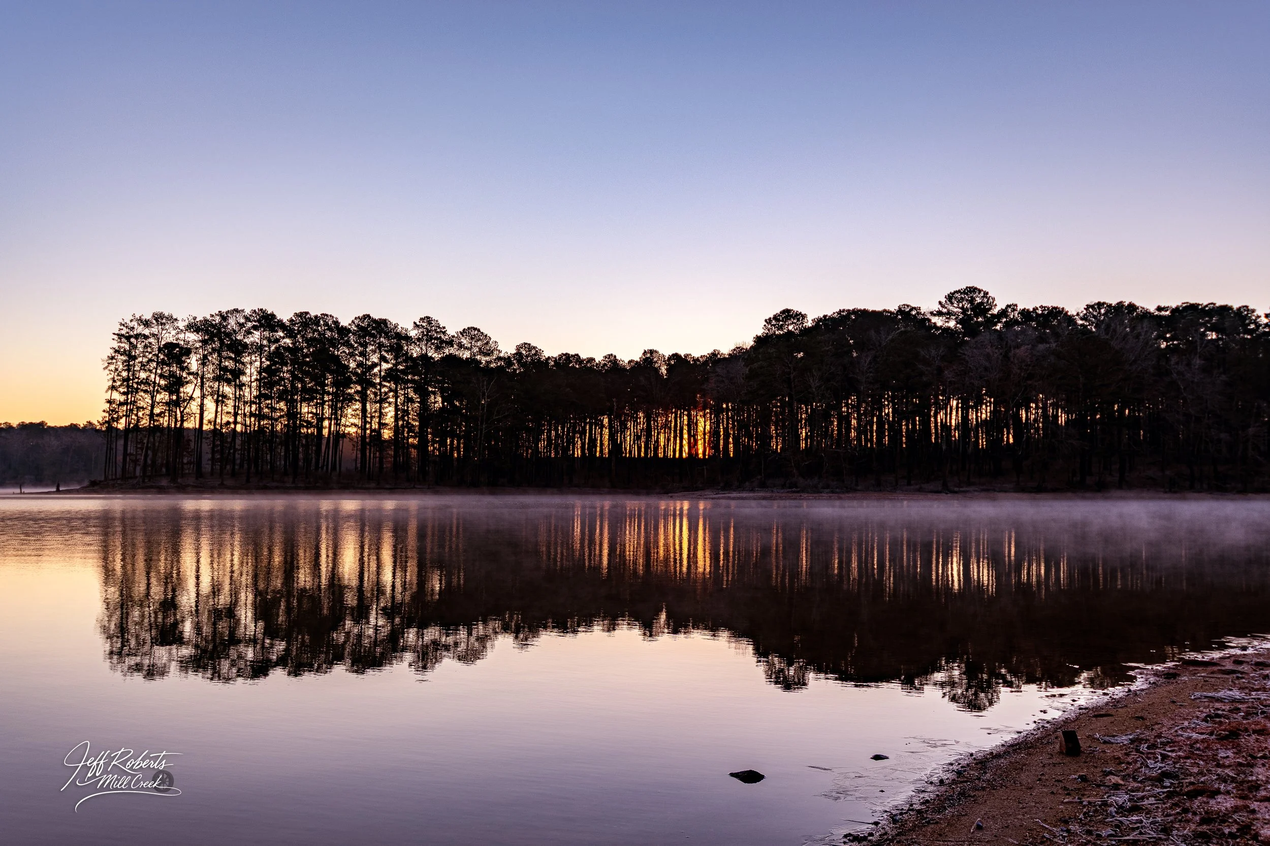 Sunset over a lake with a silhouette of a wooded shoreline reflected in the calm water, with mist rising from the surface.
