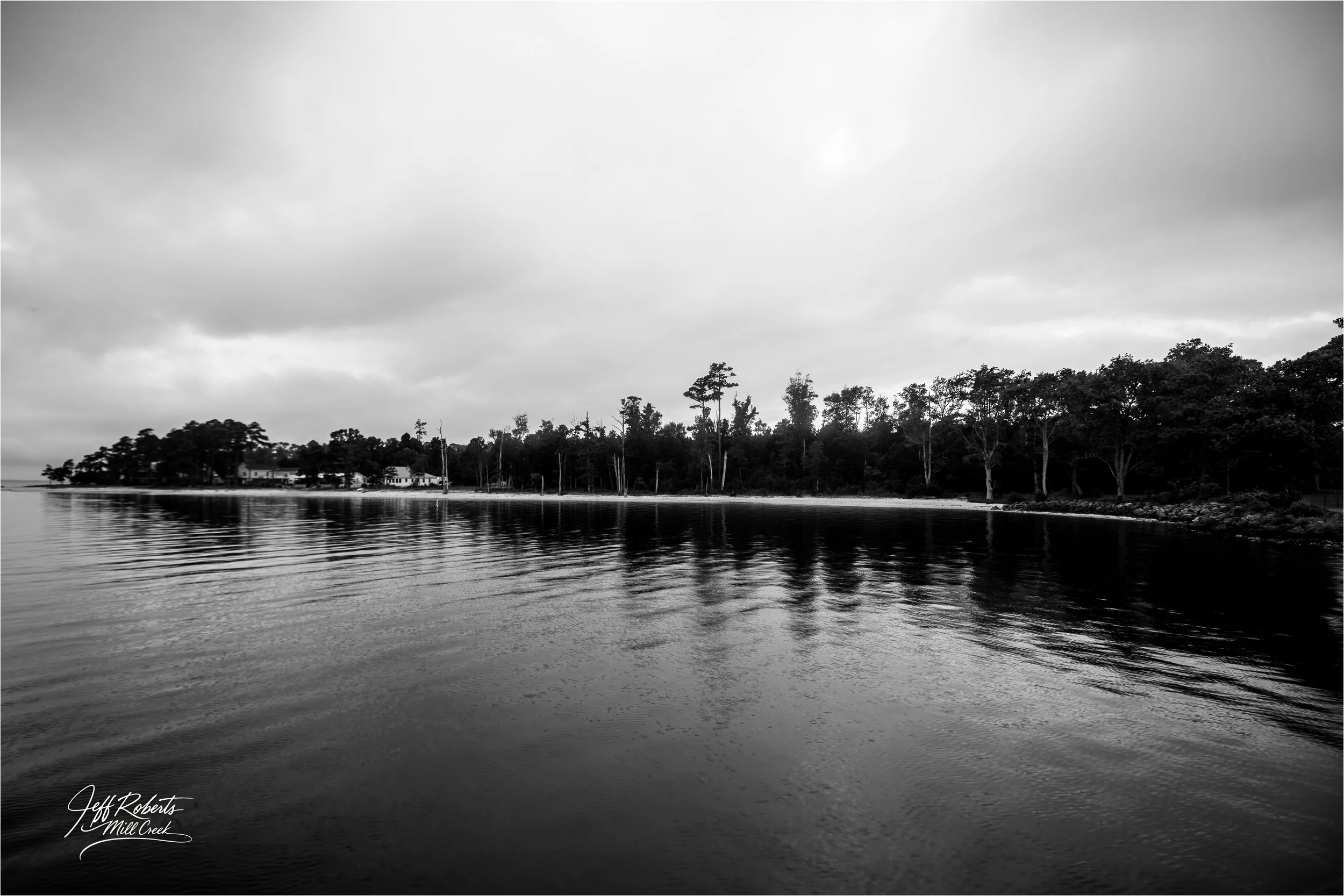 Black and white photo of calm river with reflection of trees and cloudy sky, shoreline with trees and houses in distance.