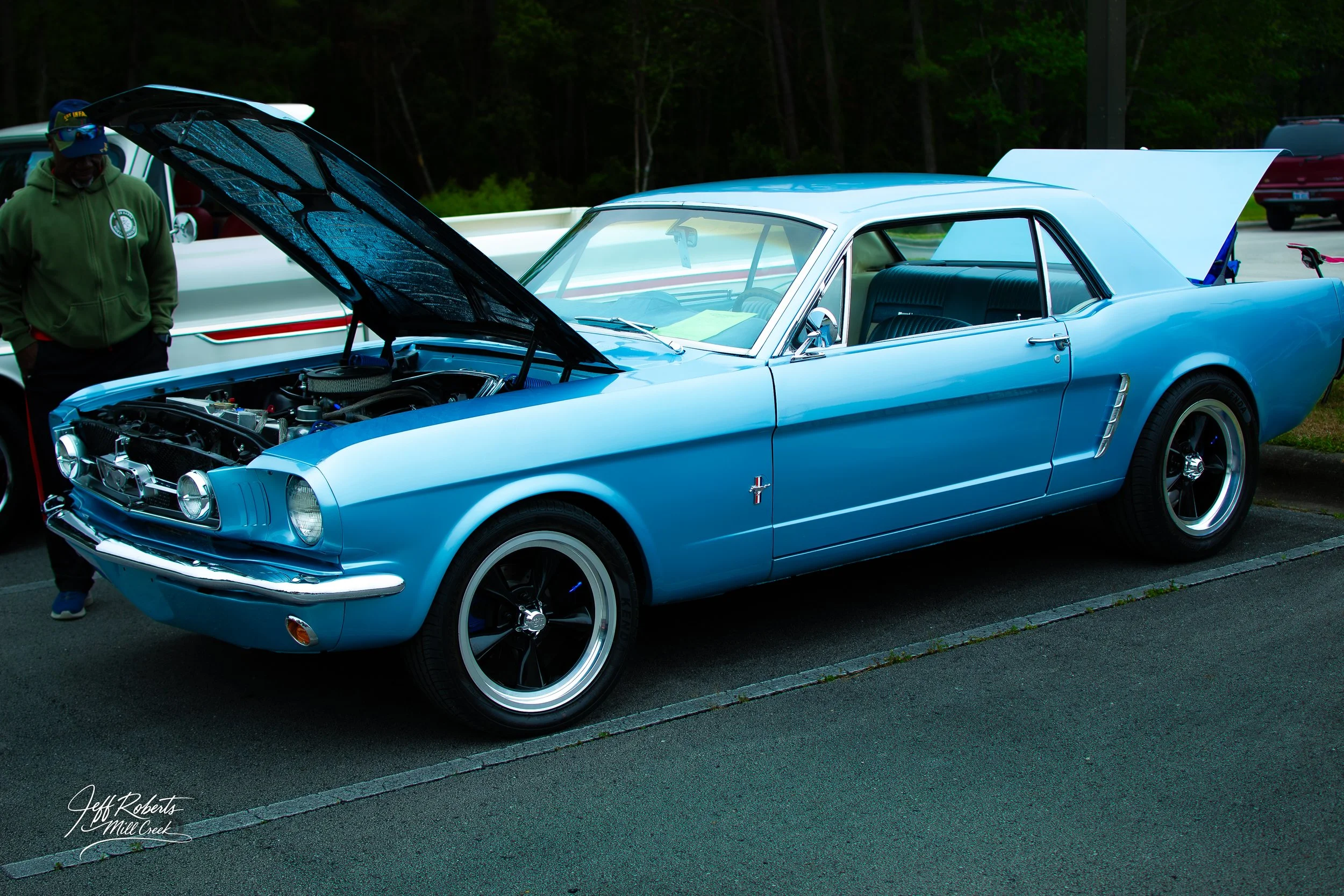 A vintage blue Ford Mustang with open hood displayed at a car show, with a person standing nearby.