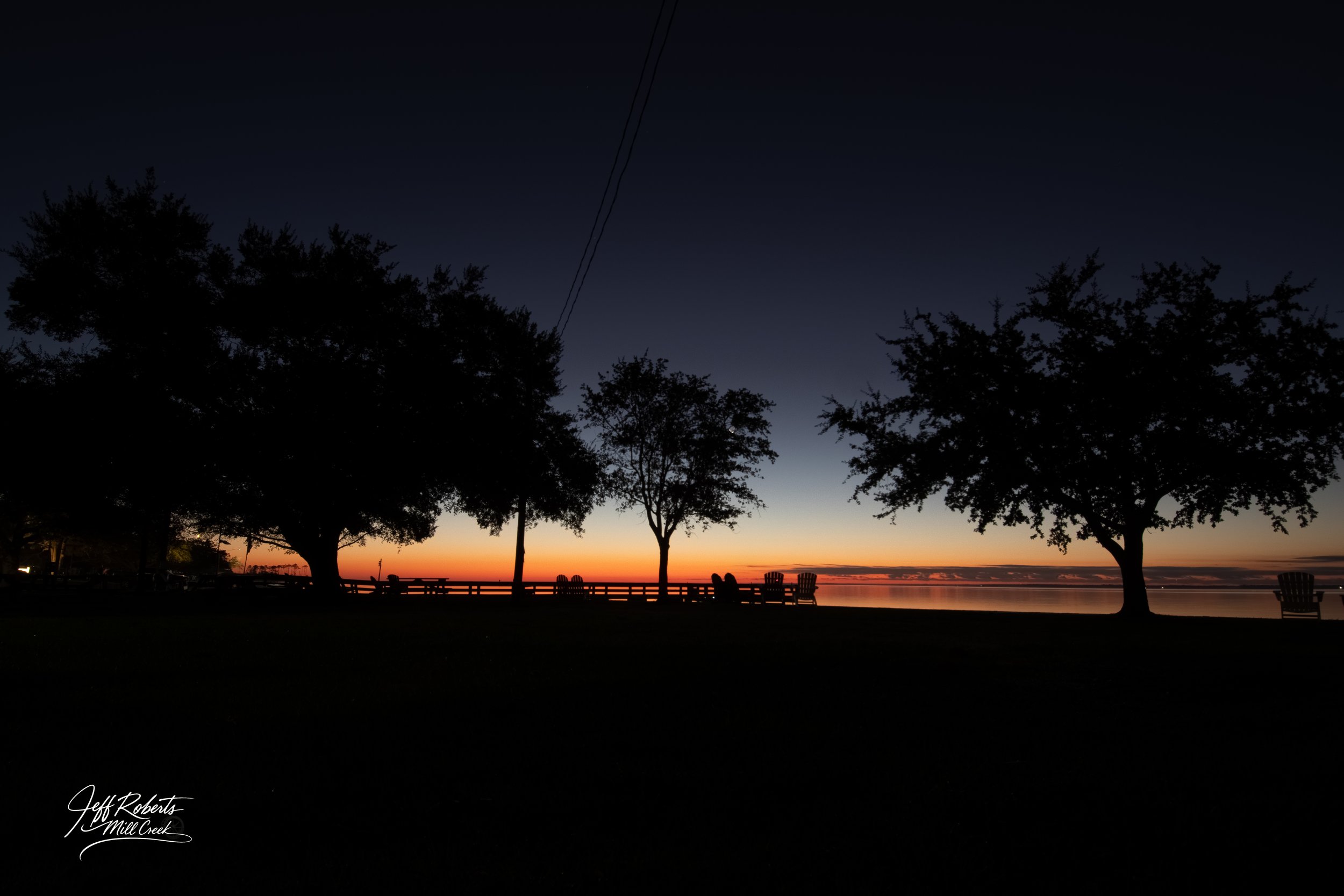 Silhouettes of trees and benches along a waterfront during sunset or sunrise, with a clear sky transitioning from orange to deep blue.