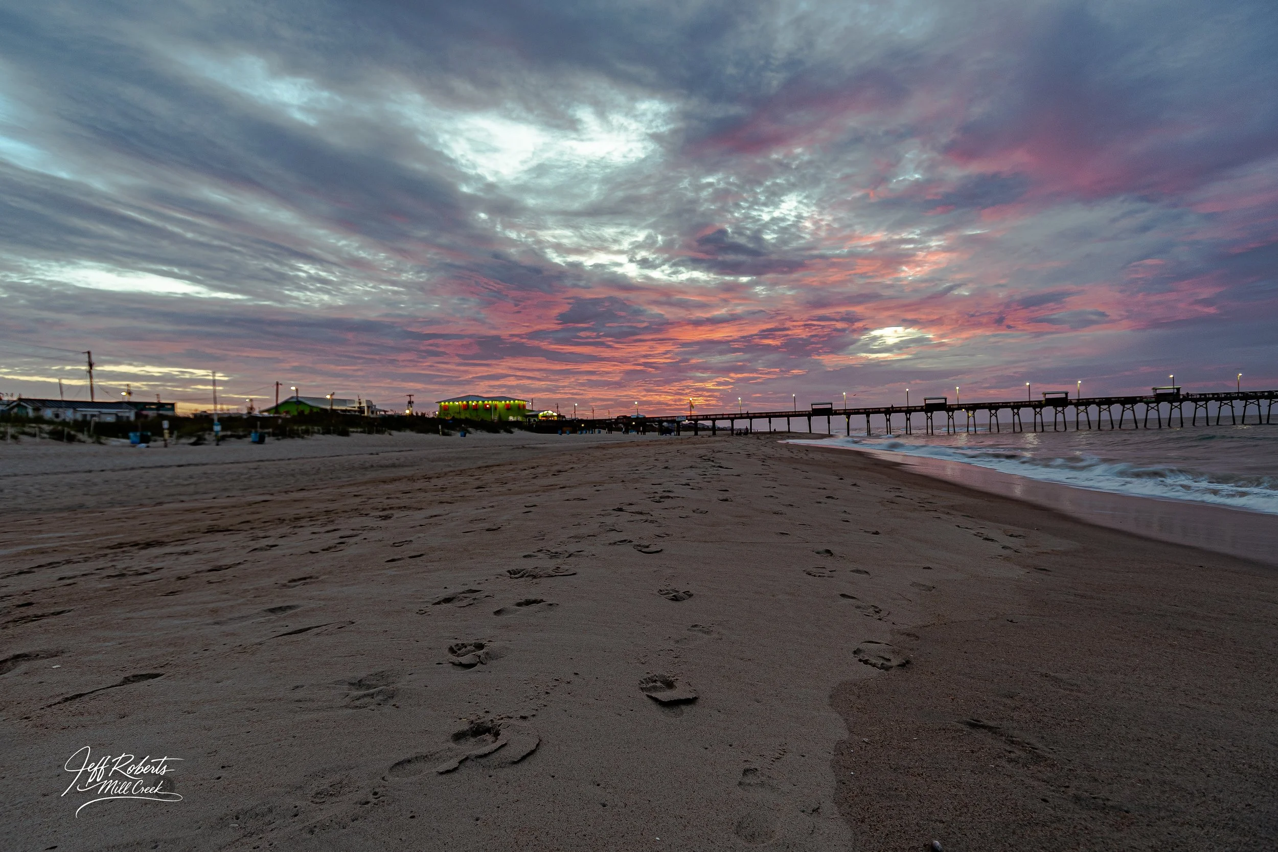 Footprints in the sand on a beach at sunset with a pier extending into the ocean and a colorful sky with pink, purple, and gray clouds.