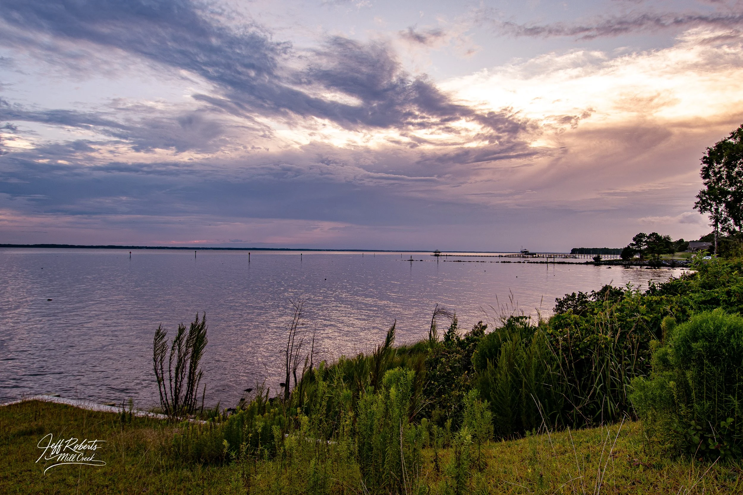 A serene body of water under a cloudy sky at sunset, with a grassy shoreline, green bushes, and distant piers.