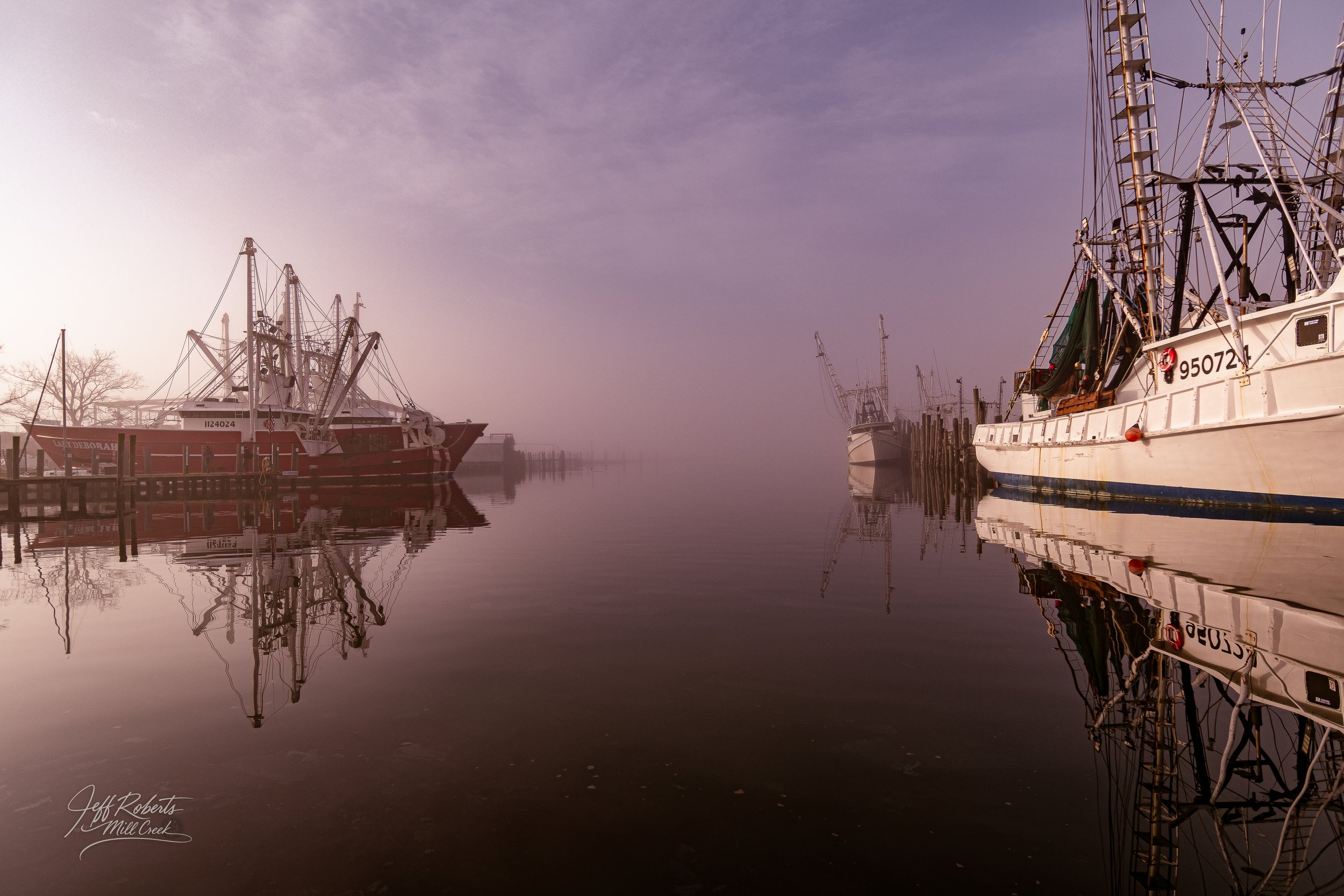 Calm harbor with fishing boats docked along the pier, reflected in still water, with a foggy, purple-hued sky overhead.