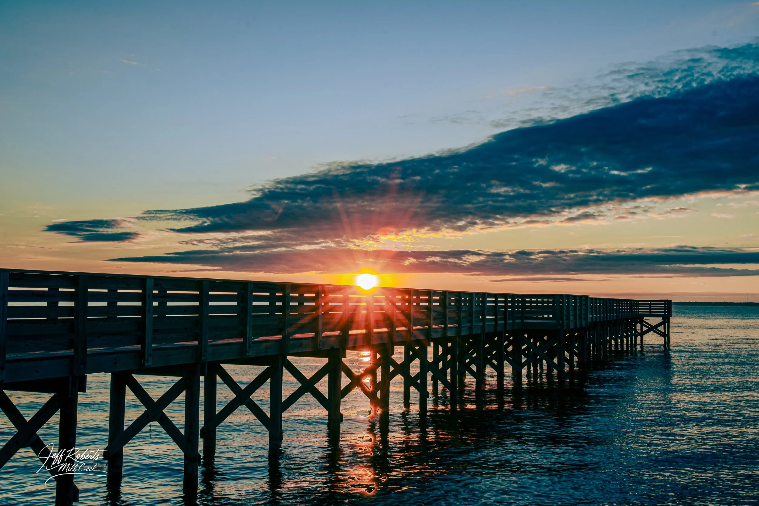 Sunset over a wooden pier extending into a body of water with clouds in the sky.