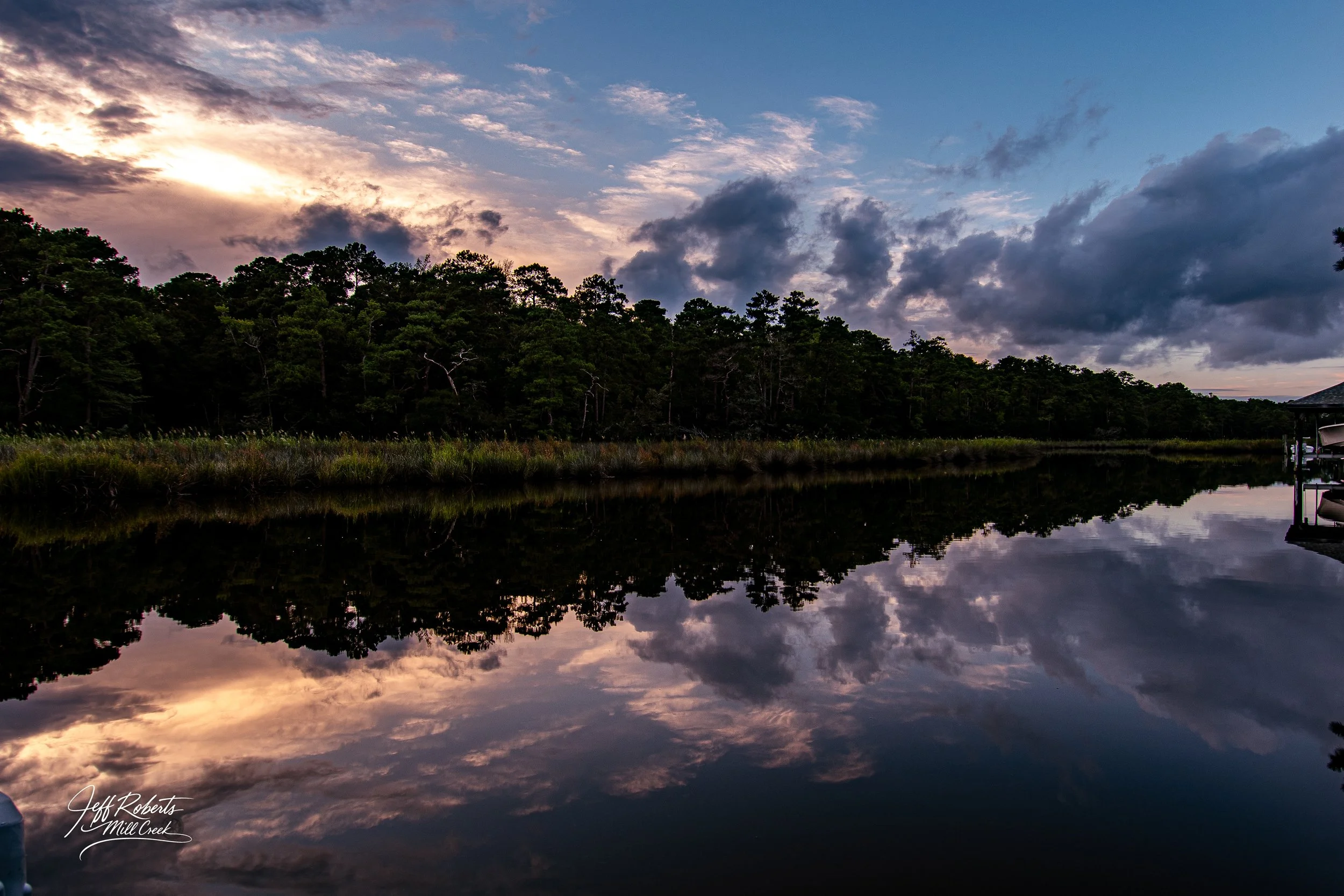 Sunset over a calm lake with a forested shoreline, sky with clouds, and their reflection in the water.