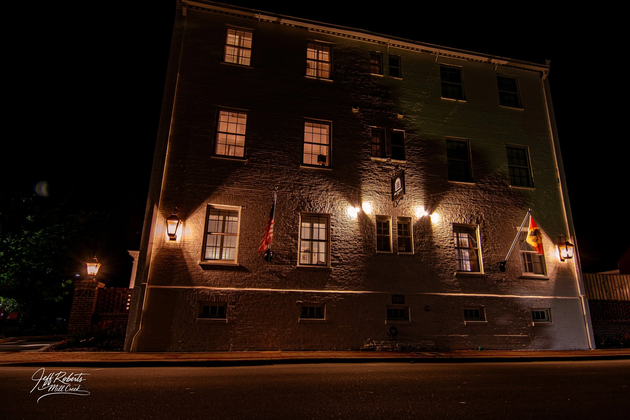 Nighttime view of an old brick building with several illuminated windows and outdoor lights, American and German flags hanging from flagpoles outside.