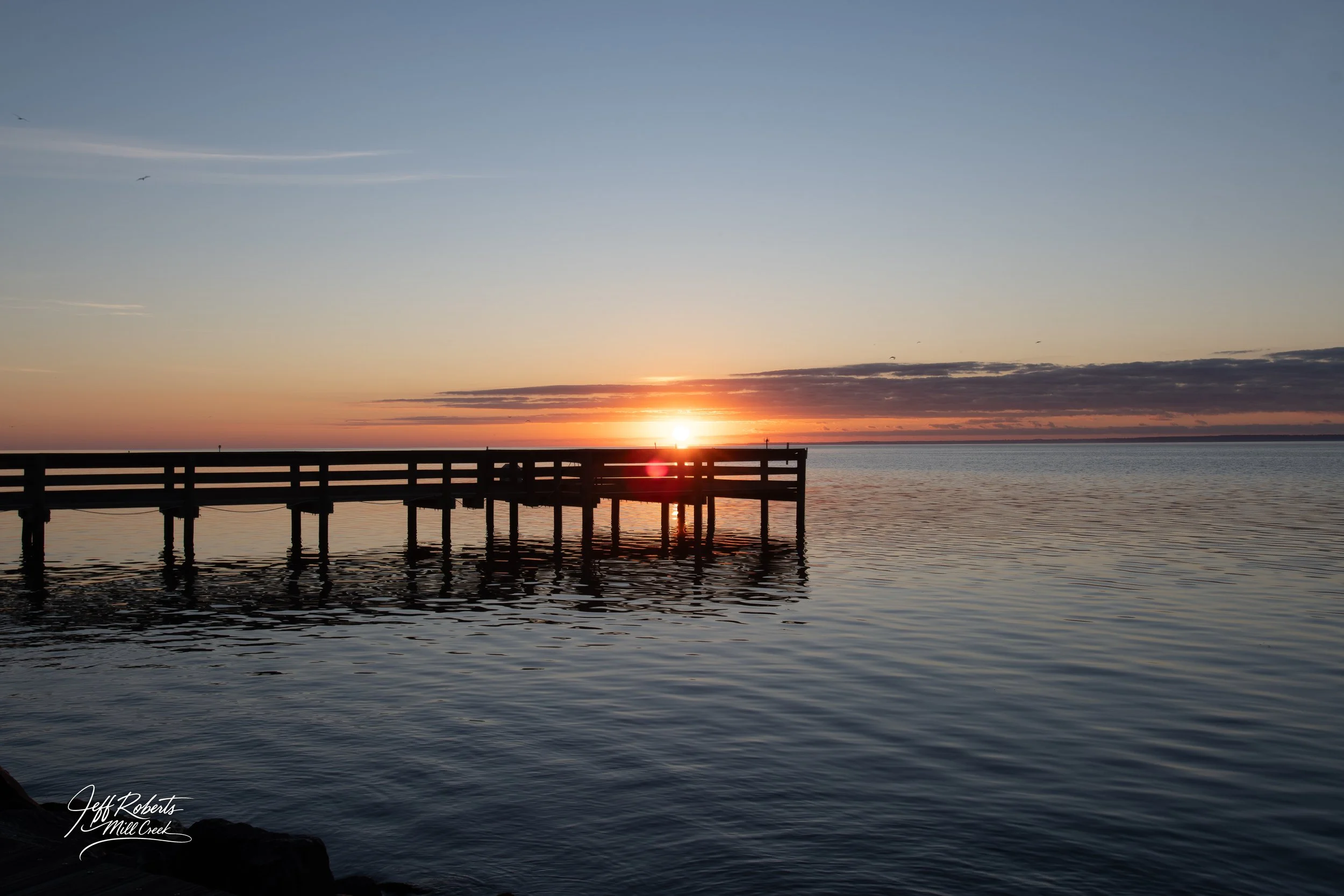 A wooden pier extending into a calm body of water at sunset, with the sun partially below the horizon and a partly cloudy sky.