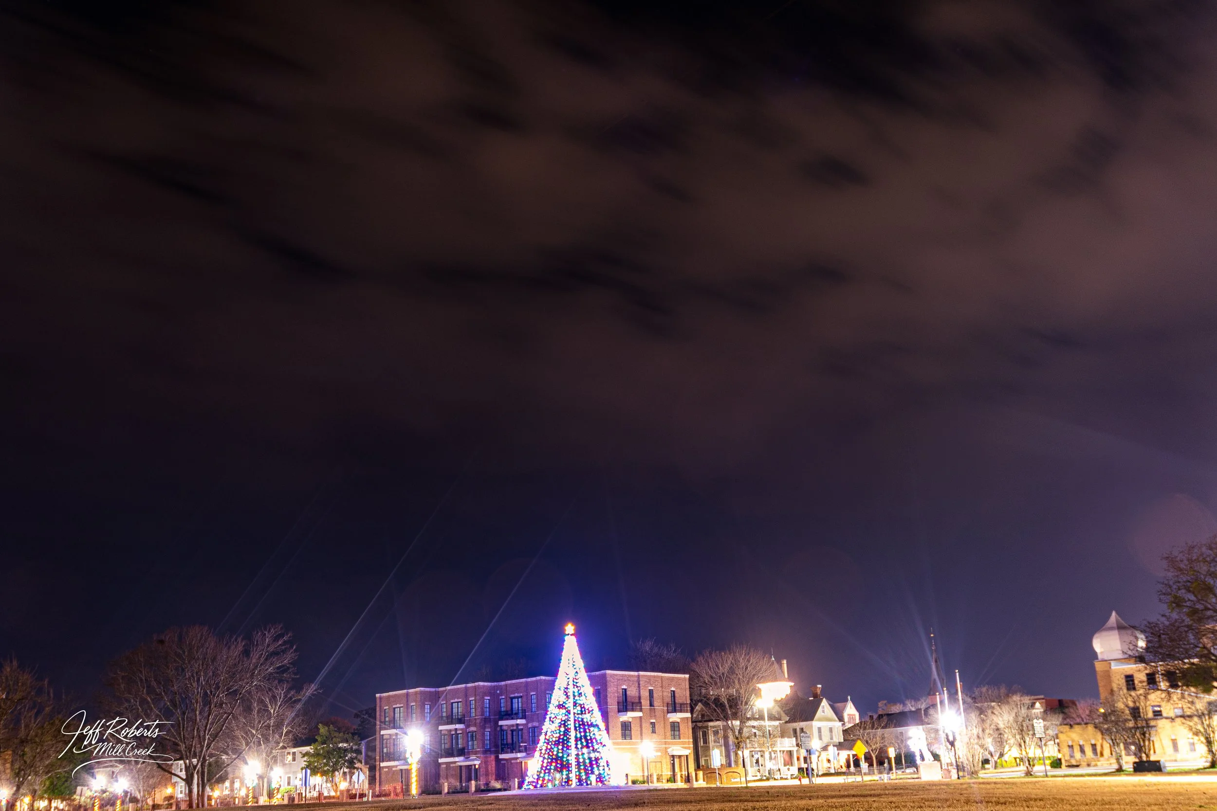 Nighttime scene in a small town with a large Christmas tree decorated with lights, and buildings with lit windows and streetlights.