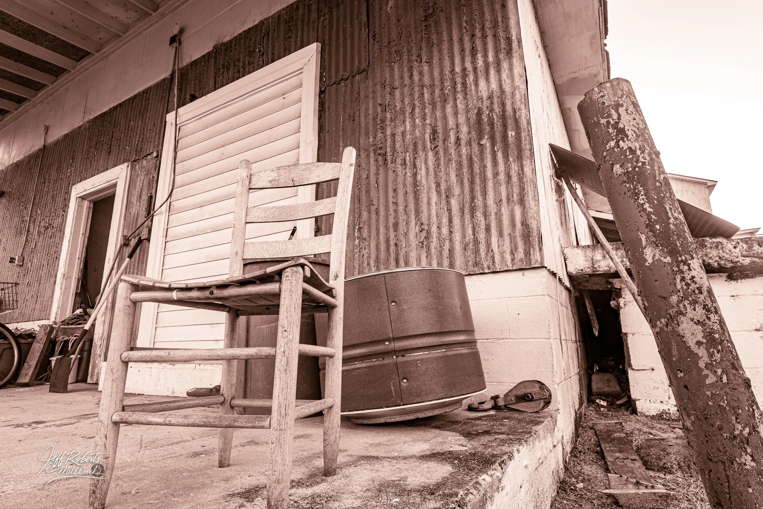 A rustic outdoor scene featuring a weathered wooden chair, a rusty barrel, and various old tools and objects against a corrugated metal wall with peeling paint.
